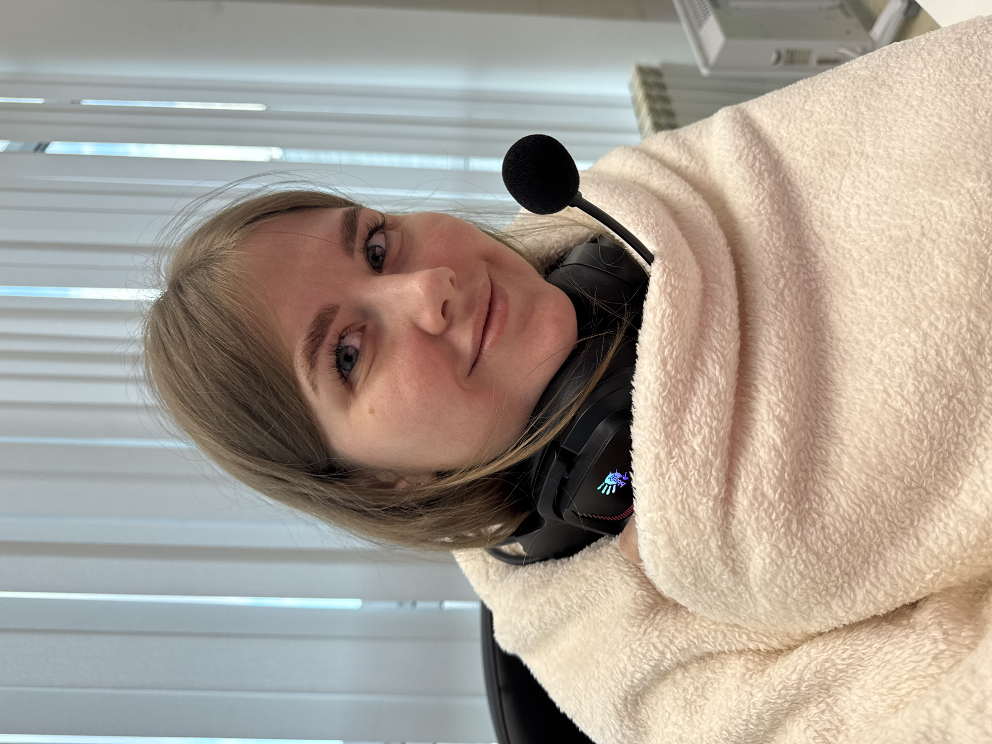 A fair-skinned woman with light brown hair is wrapped in a fluffy, light beige blanket. She is wearing black headphones with a microphone and a colorful logo on the earcups. The background consists of white window blinds and office equipment, indicating an indoor office environment. It is not possible to determine the location as Perm, Russia, from the image.