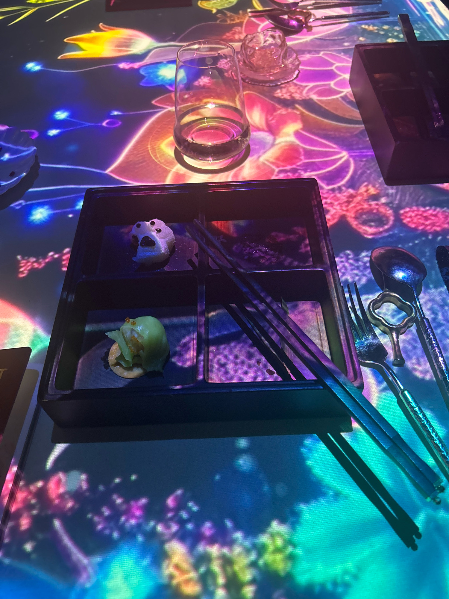The image depicts a close-up, high-angle view of a dining setting on a table illuminated by a vibrant projection of colorful, abstract floral patterns, likely in Shanghai, China. In the foreground, a black bento-style dish holds two small appetizers. The upper compartment contains a white, circular item with black specks, resembling a small dumpling or dessert. The lower compartment holds a light green, possibly steamed, dumpling with a few orange pearls on top, resting on a circular base. A pair of dark chopsticks rests diagonally across the bento box. To the right of the bento box, a glass of clear liquid is visible, partially obscured by the projections. Further to the right, a set of silverware, including a fork and spoon, is laid out, with the spoon featuring an ornate, flower-shaped handle. The background is dominated by the dynamic light projections, creating a surreal and immersive dining environment. The overall scene suggests a modern, artistic dining experience, potentially in a restaurant that utilizes interactive projections for ambiance.