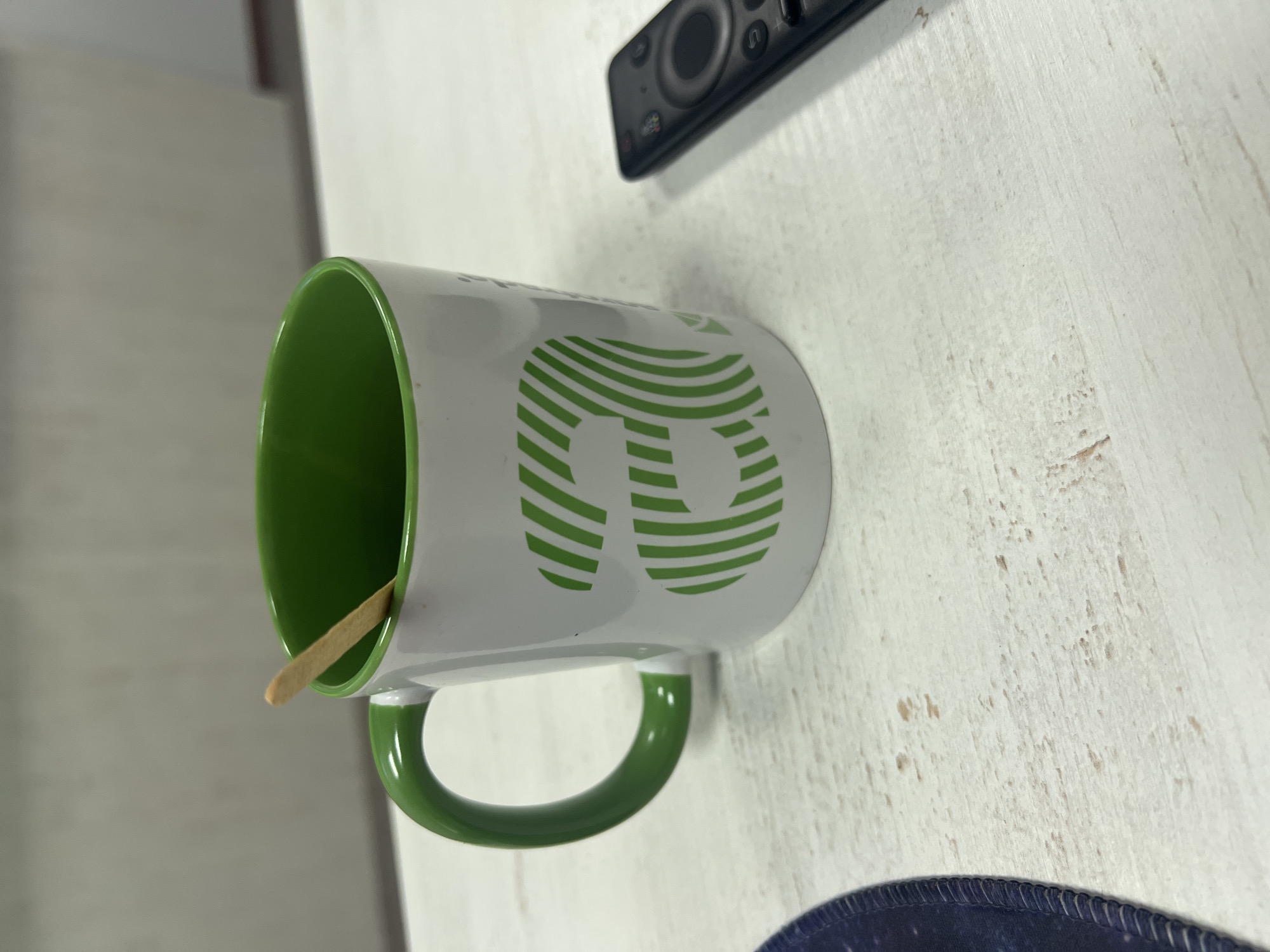 The image shows a white mug with a green interior and handle. The mug has a green striped "B" on its side and contains a wooden stirrer. The mug is sitting on a white surface, possibly a table. A black remote control is partially visible above the mug, and part of a dark blue circular object, possibly a mousepad, is visible below it. The location is unknown, and there is no evidence to suggest it is in Poltava, Ukraine. No people, actions, or interactions are visible.