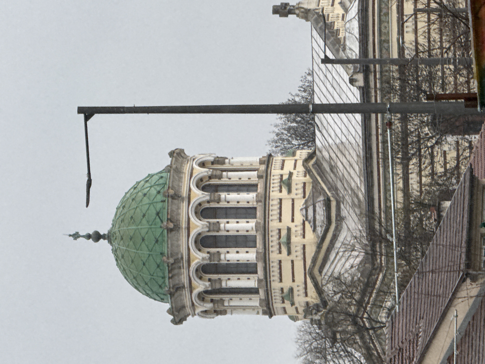 The image shows the dome of the Sofia Synagogue, located in Sofia, Bulgaria. The green, patterned dome sits atop a multi-tiered, ornate building section with arched windows. A streetlight pole with a horizontal arm extends across the left side of the frame. The rest of the building is visible to the right. Branches from bare trees are visible in the background. The sky is overcast and gray. Tiled roofing is visible on the lower right side of the frame.