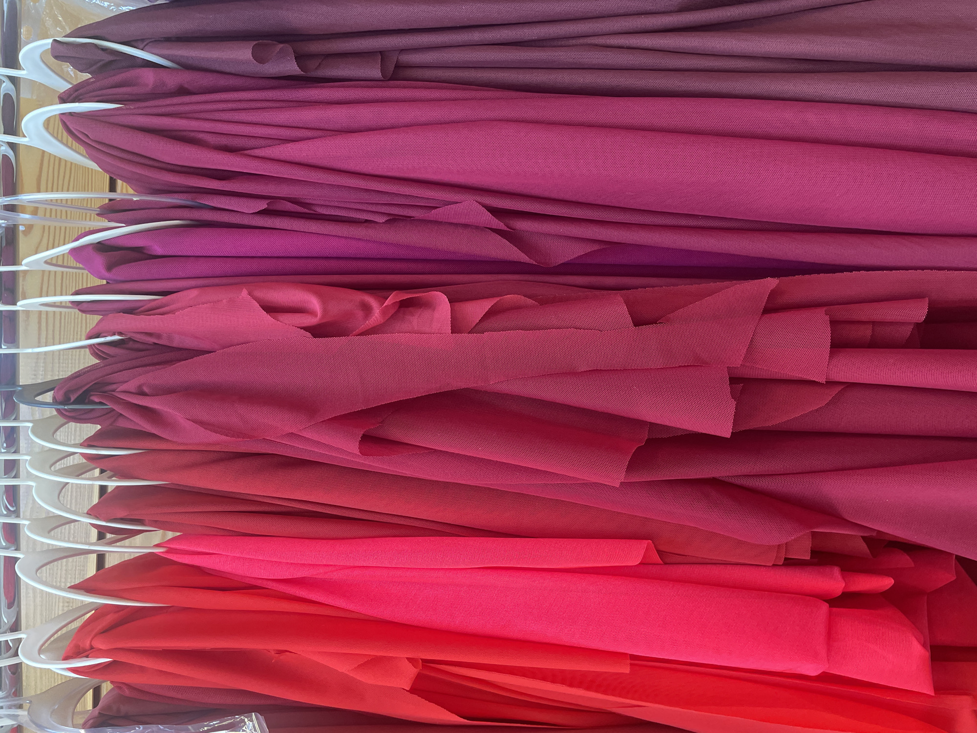 This is a close-up, eye-level, indoor shot of several pieces of fabric hanging on white hangers on a rack. The fabric colors range from bright red on the left to deep burgundy on the right, with various shades of pink and magenta in between. The fabric appears to be lightweight and draped, with some pieces having a ruffled texture. The background is out of focus and appears to be wooden slats, suggesting the rack is in a closet or storage area. There are no people or animals present in the image.