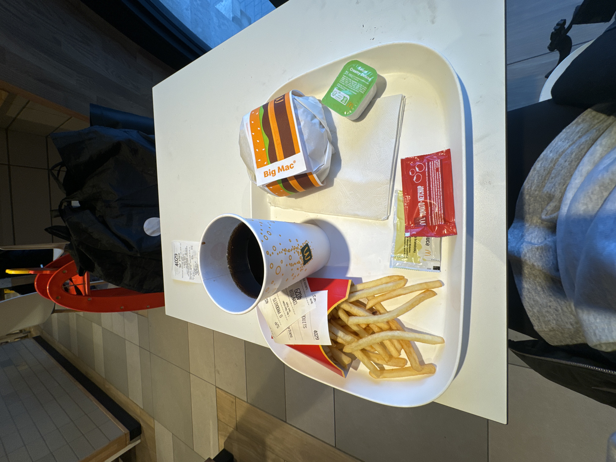 The image shows a McDonald's meal tray on a white table, containing a Big Mac, french fries, a drink (likely Coca-Cola), dipping sauce (Creamy Deluxe), and ketchup. The tray also has receipts. The location is likely in Paris, France based on the "Sur place" on the receipt, which is French for "on the spot" and usually appears in McDonald's receipts in France. There is a black bag and a red object in the background. The environment includes a tiled floor and a window, suggesting an indoor seating area. A portion of a person, possibly seated, is visible on the right side of the frame.