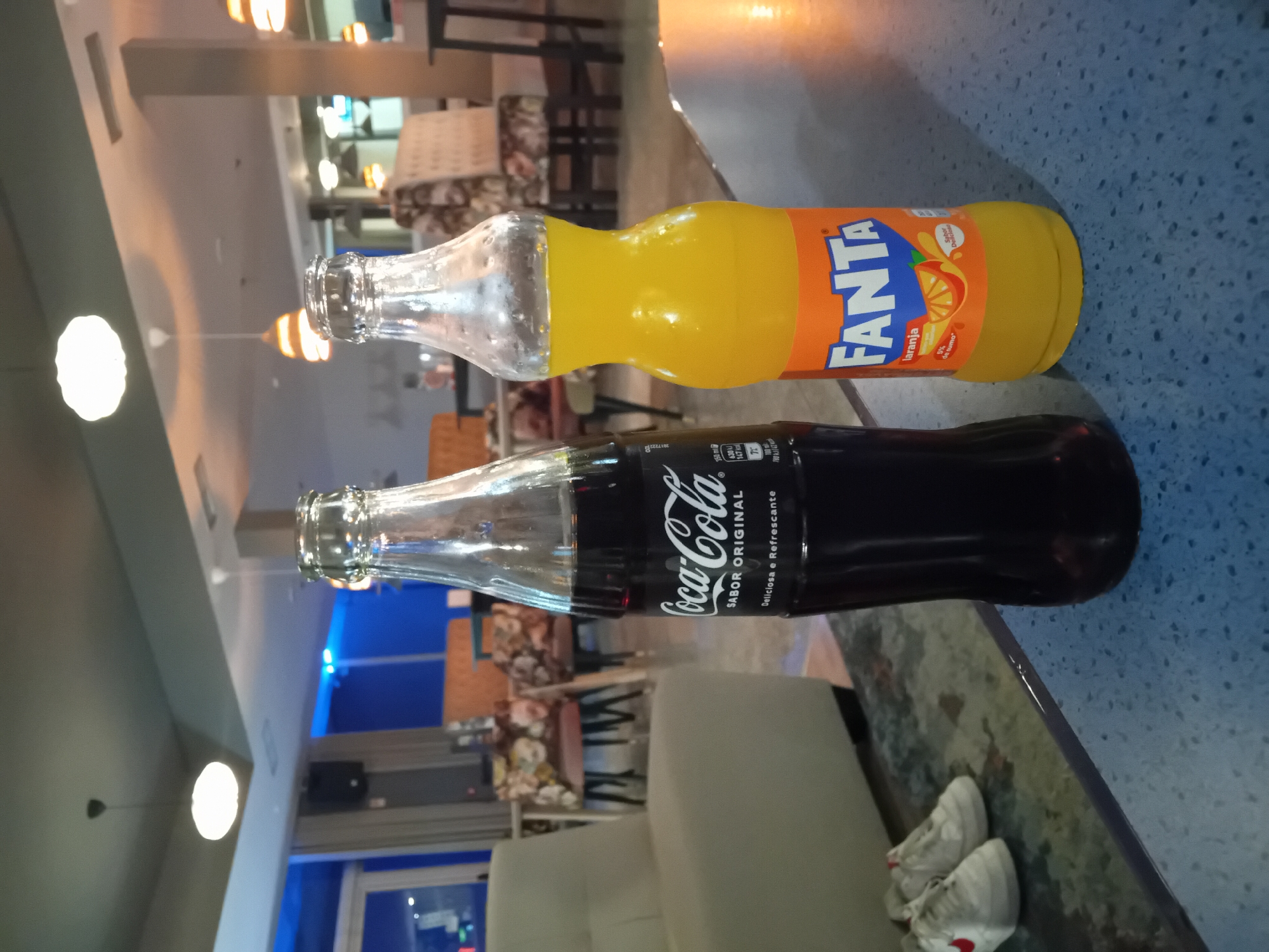 The image shows two glass bottles, one containing Coca-Cola and the other Fanta orange soda, lying on a table in an indoor setting. The Fanta bottle is filled with orange liquid and displays the Fanta logo. The Coca-Cola bottle contains a dark liquid with the "Coca-Cola" label printed on its side. The background reveals a seating area with chairs and lighting fixtures. Two pairs of white athletic shoes are at the edge of the table.