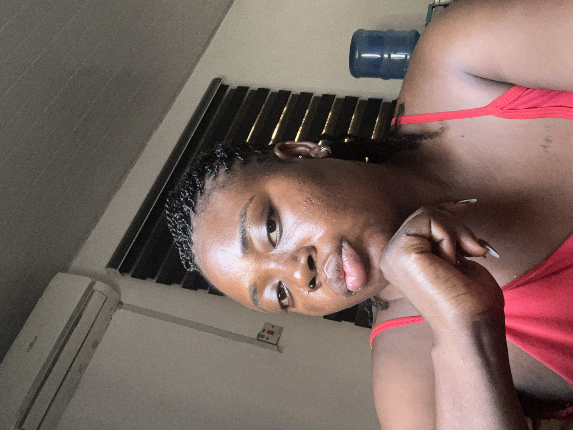 This is a portrait of a woman indoors, with the camera angled from above. She has dark skin and her hair is styled in braids. She is wearing a red top. Her left hand is resting on her chin, and her right arm is visible. The background includes a dark-slatted blind, an air conditioning unit, and a large blue water dispenser bottle. The lighting suggests it is daytime, possibly late afternoon or early evening, as there are some shadows. The scene appears to be in a residential setting. The woman's expression is neutral. There is a power outlet visible on the wall.