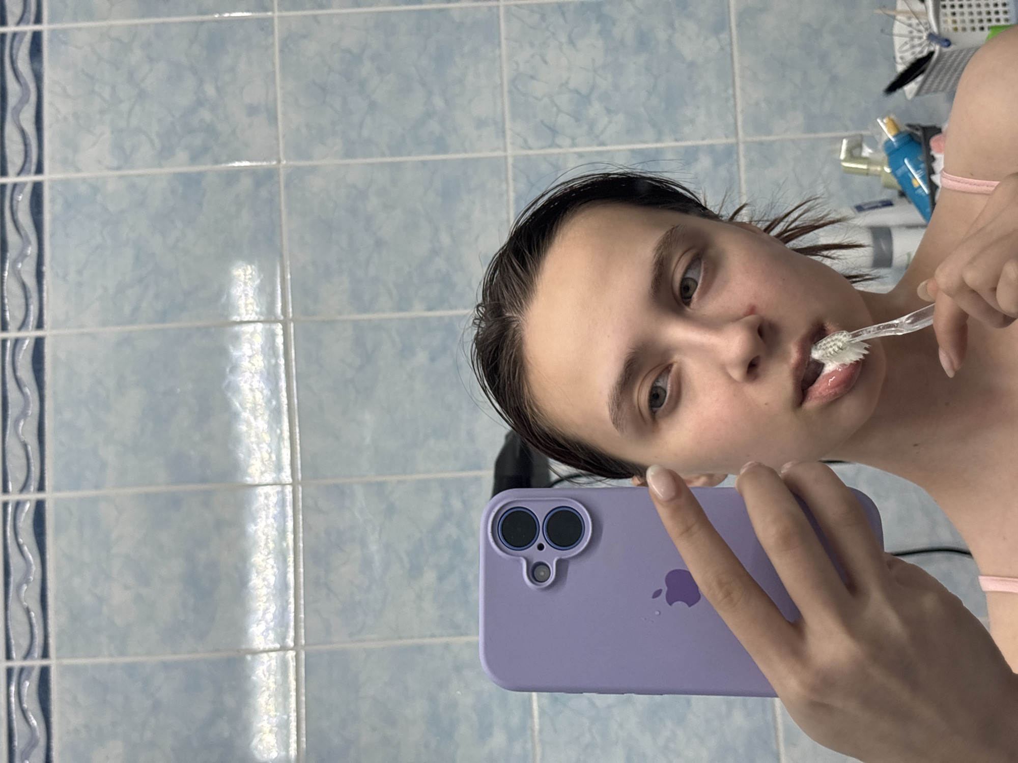 The image shows a young woman brushing her teeth in a bathroom. She is holding a light purple iPhone in her left hand and a clear toothbrush in her right hand, with white toothpaste on the bristles. The woman has wet, dark hair and light skin. The background consists of blue and white tiled walls. Other bathroom items are visible in the upper right corner, including tubes, containers, and a toothbrush holder. The location appears to be a private residence, though specific information regarding the city of Прикамский, Russia, cannot be verified based on the image alone.