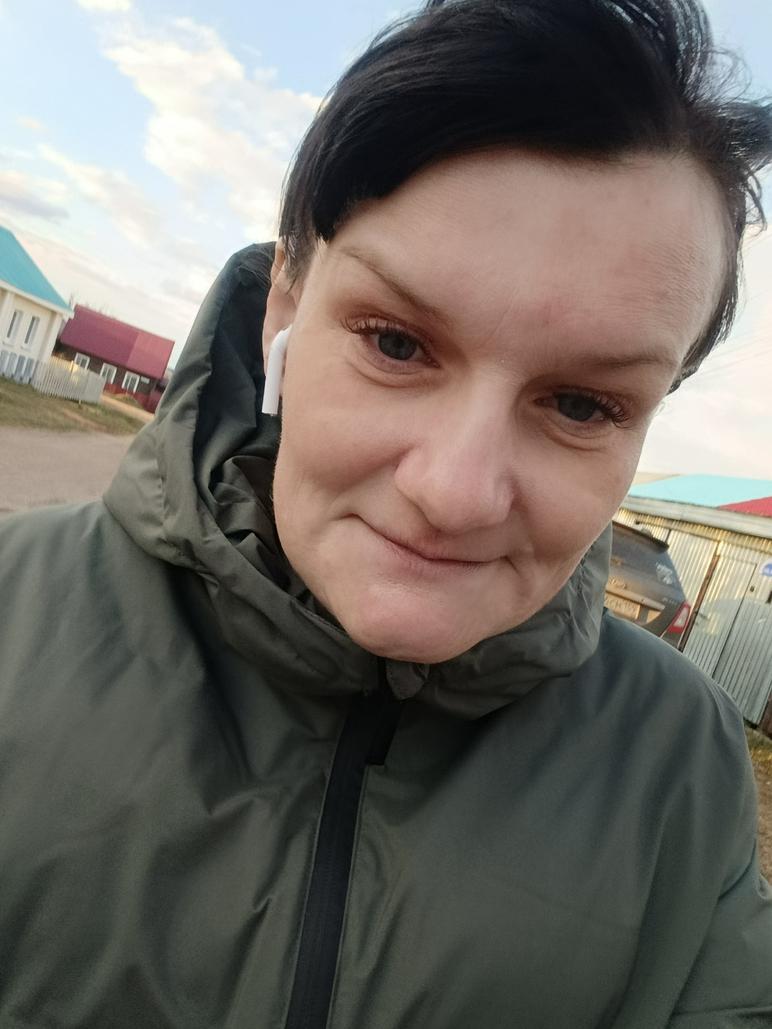 A woman is captured in a close-up, slightly low-angle selfie outdoors in what appears to be a residential area in Фоки, Russia. She has dark hair and is wearing a dark olive green hooded jacket with a black zipper. A white earbud is visible in her right ear. Her expression is calm and she has a gentle, subtle smile, looking directly towards the camera.
The background reveals a neighborhood setting with houses under a bright sky with scattered white clouds. To the left, a white house with a light blue roof is partially visible, and further back, a building with a red roof is seen behind a white picket fence. On the right, a corrugated metal fence or building is present, with a dark car partially visible behind it. The car's license plate has blurry characters that are not legible. The lighting suggests it is either late afternoon or early evening, with soft, natural illumination. No clearly readable text is visible in the image.