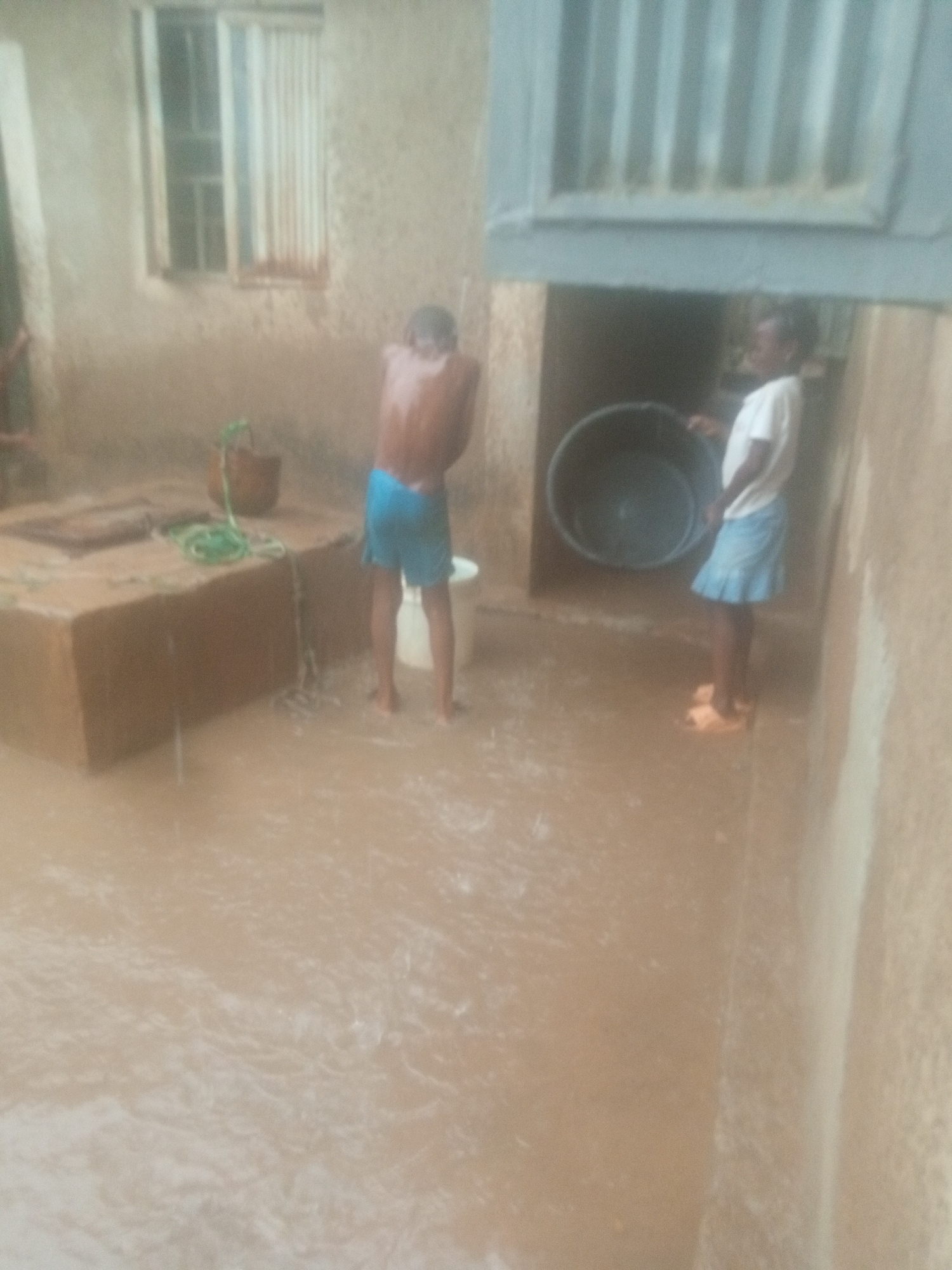 The image shows a flooded outdoor area in Unknown, Unknown. The water level is high enough to submerge part of a concrete structure, and a window is visible on a nearby building. Two children are standing in the water; one is shirtless and wearing shorts, while the other is wearing a t-shirt and skirt and holding a large black tub.