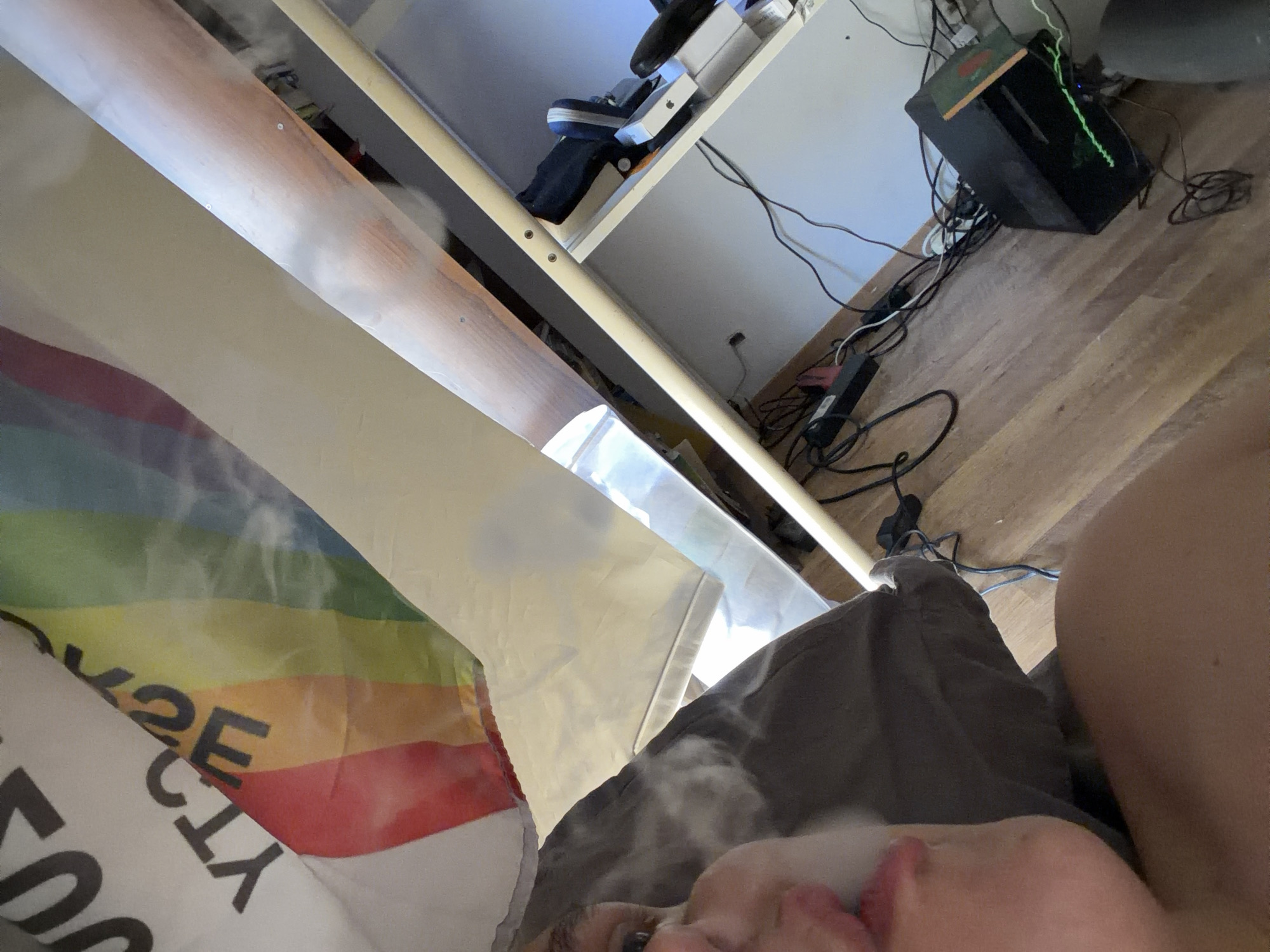 The image depicts a close-up, low-angle view of a person exhaling vapor in front of a rainbow flag. The flag appears to be partially draped over something, obscuring some of its pattern. To the right of the flag, a shelf or desk area is visible, containing electronic devices, cables, and what appears to be a computer tower. The background consists of a wall and a wooden floor. The scene is indoors, and the lighting suggests it might be daytime, though artificial light sources are also present. There is no visible indication of the specific location being Germasogeia Municipality, Cyprus; this information is outside the scope of what can be determined from the visual content alone.