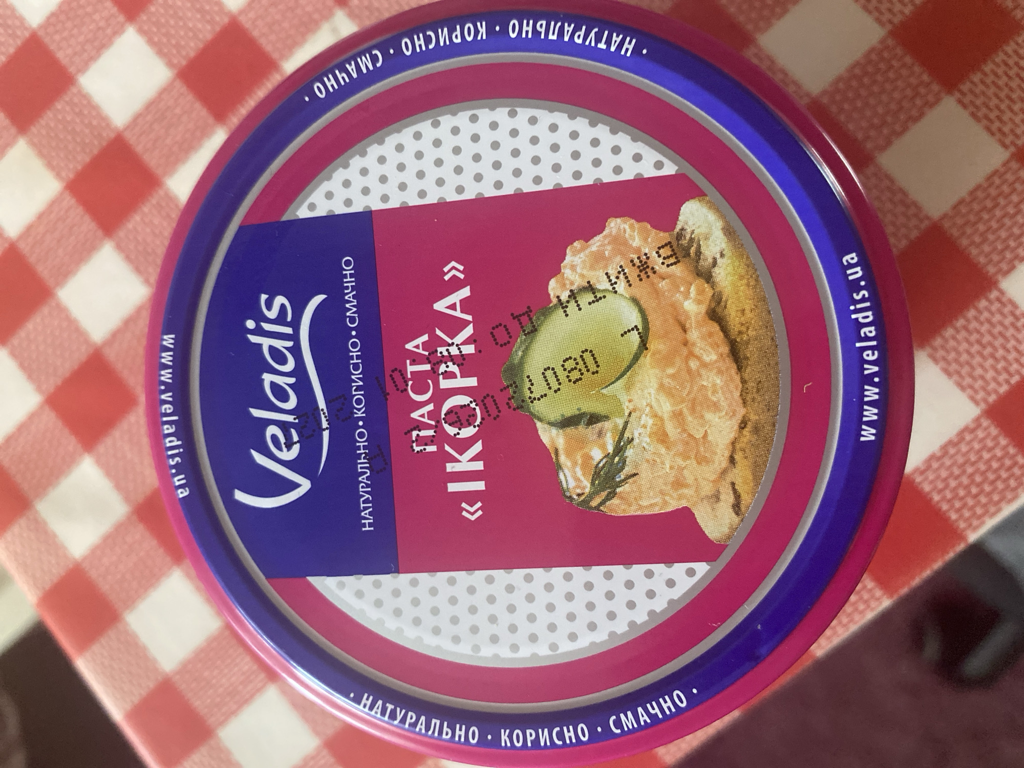 The image shows a round, metallic lid of a food product, likely a spread or paste, placed on a red and white checkered tablecloth. The lid features a vibrant pink and purple design. Prominently displayed are the brand name "Veladis" in a stylized white font, and the product name "ПАСТА «ІКОРКА»" in a similar font. Surrounding the central design are words in Ukrainian: "НАТУРАЛЬНО • КОРИСНО • СМАЧНО" (Natural • Healthy • Tasty) and the website "www.veladis.ua". A small, slightly blurry image of the food product itself, a creamy orange spread with a sprig of dill and a slice of cucumber, is visible in the center of the lid. The lighting suggests an indoor environment, possibly a kitchen or dining area. No specific location in the city of Zhytomyr, Ukraine, can be identified from the provided image.