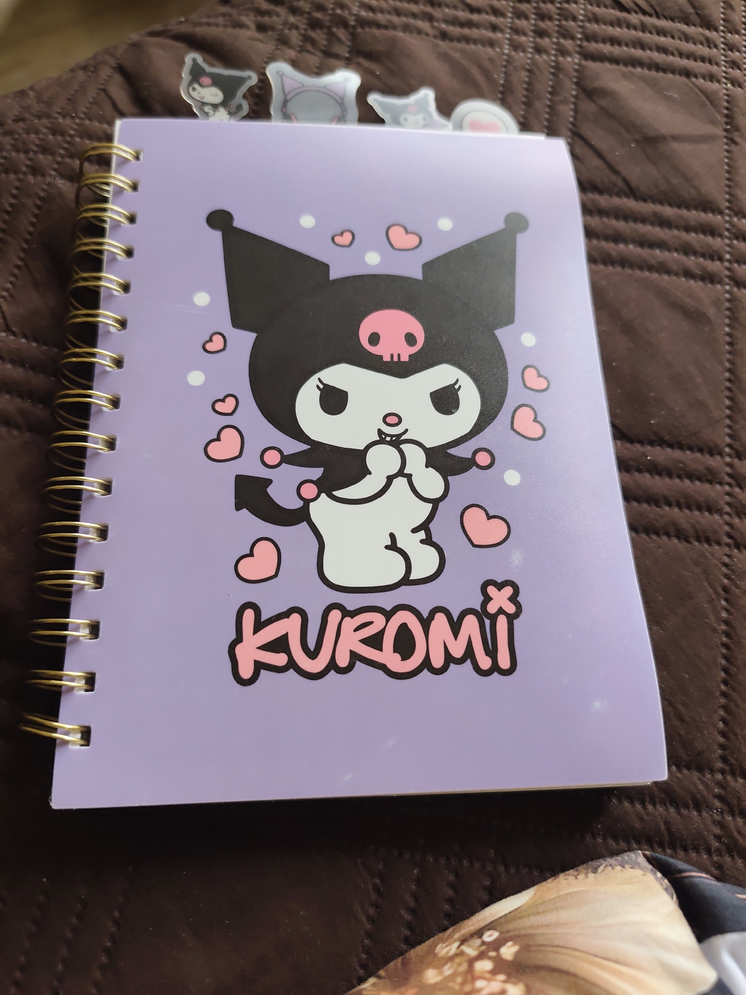 The image shows a spiral-bound notebook with a purple cover. The cover features a cartoon drawing of the character Kuromi, surrounded by hearts and polka dots. The word "KUROMI" is printed below the character. Several small character bookmarks are visible sticking out of the top of the notebook. The notebook sits on a textured brown surface, likely a blanket or upholstery. The location is not identifiable from the given image.