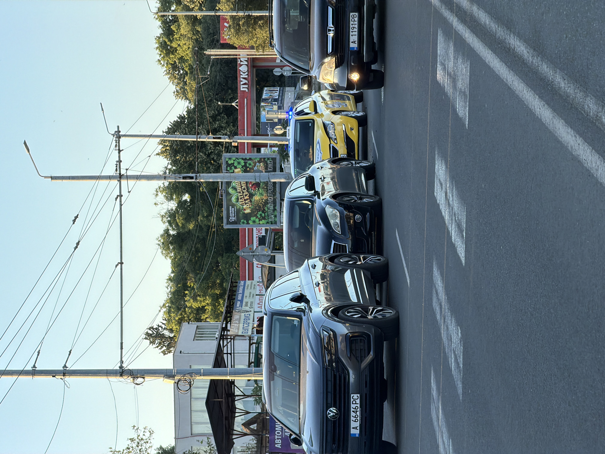 The image shows a street scene in Burgas, Bulgaria, with several cars stopped at a crosswalk. In the foreground, a dark grey Volkswagen Touareg is parked. To its right, a black sedan is partially visible, followed by a yellow taxi with blue lights on its roof. A black Honda CR-V is to the right of the taxi. In the background, there are buildings, trees, streetlights, and overhead wires. A "Lukoil" gas station sign is visible in the distance. The sky is clear and blue.
