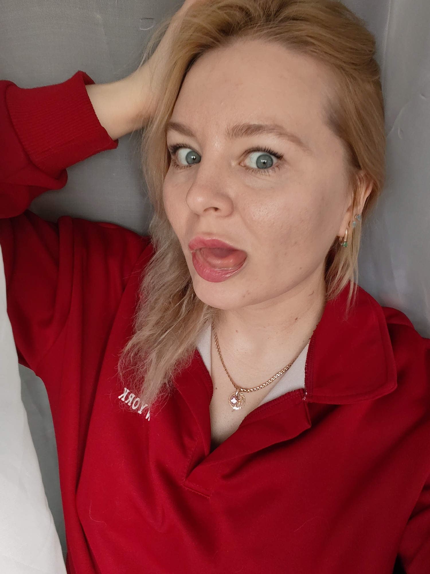 A light-skinned woman with blonde hair and blue eyes is visible in the image. She is wearing a red collared shirt with white lettering that is mirrored and illegible in the photo. She has her mouth open, exposing her tongue. She has gold hoop earrings and a gold necklace with a pendant. Her left hand is raised, fingers touching her hair. The background is a plain gray surface. The location is unspecified and there is no information to suggest that this picture was taken in the city of Izhevsk, Russia.