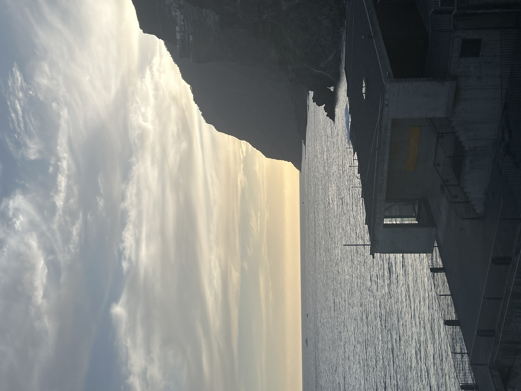 The image shows a coastal view in Câmara de Lobos, Portugal, at what appears to be dusk or dawn. On the left is a sky filled with cirrus clouds, transitioning to a golden hue near the horizon. A dark, mountainous coastline follows. To the right is the ocean, with a building or structure extending out. The structure has a concrete appearance and is adjacent to the ocean.