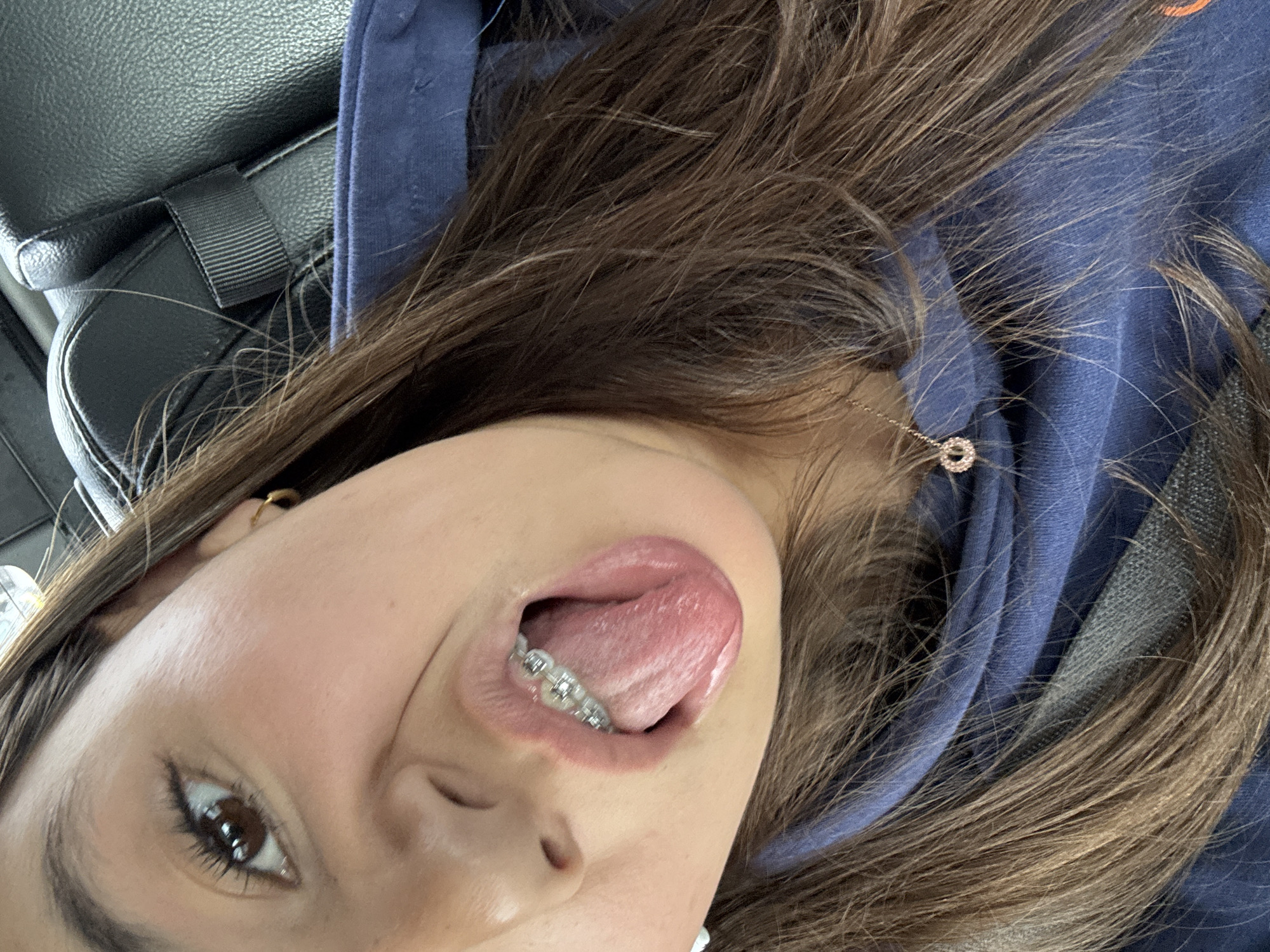 A close-up selfie shows a young woman lying down, possibly in a car given the presence of a seatbelt in the upper left corner. She has brown hair and is wearing a dark blue hoodie. Her tongue is sticking out, revealing braces on her lower teeth. The background is blurry, suggesting the focus is primarily on her face. The location, Austin, United States of America, is not discernible from the image alone.