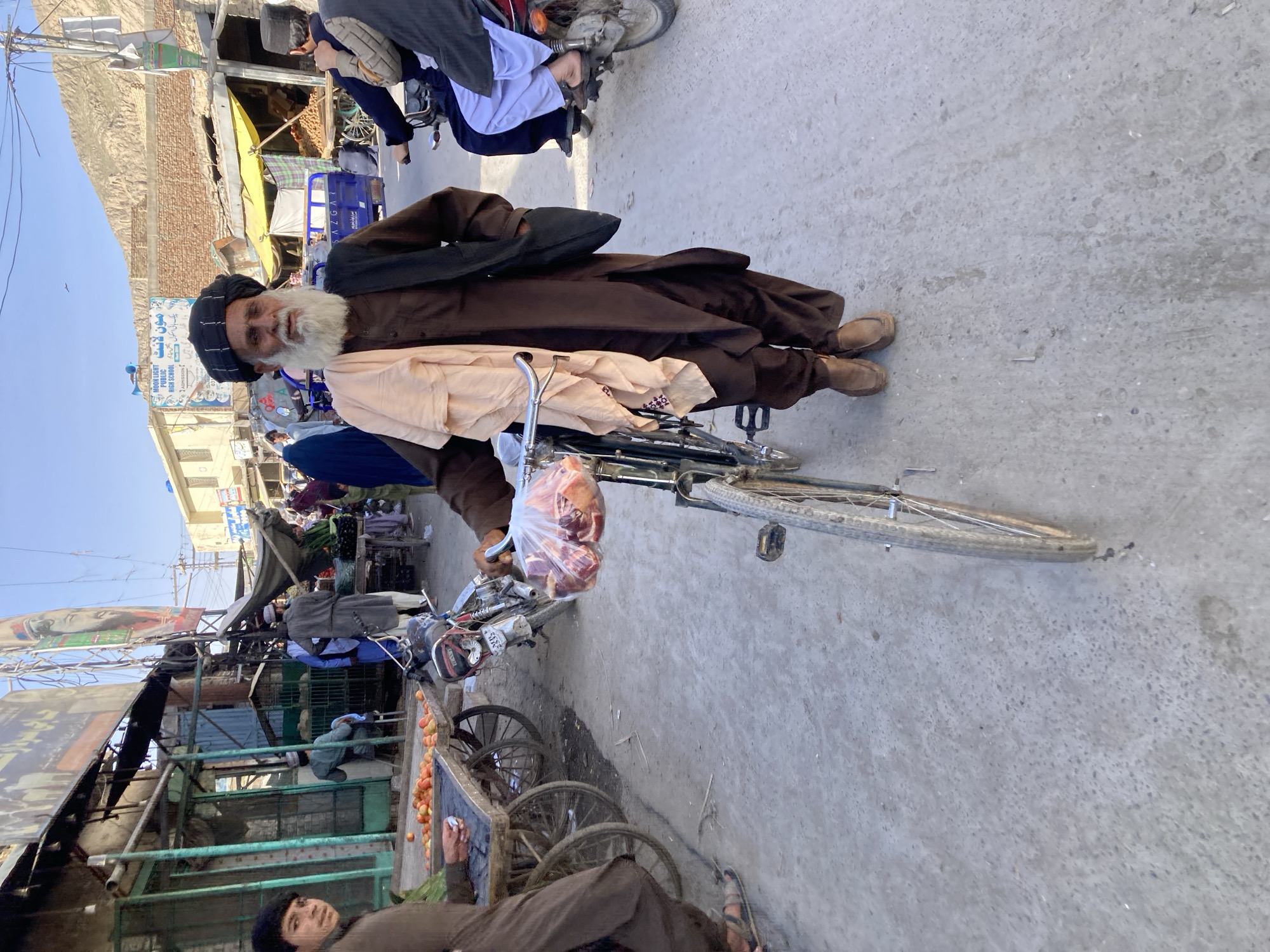 A wide-angle photograph depicts a man in traditional attire standing next to a bicycle on a street in Quetta City Tehsil, Pakistan. The man wears a dark head covering, a long-sleeved brown shirt, brown pants, and brown shoes. He has a long white beard. He stands next to a bicycle with a package in his hand. Other people are visible in the background, including people on motorcycles. There are vendor stalls and buildings along the street. Overhead power lines are visible. The ground is paved and appears dusty.