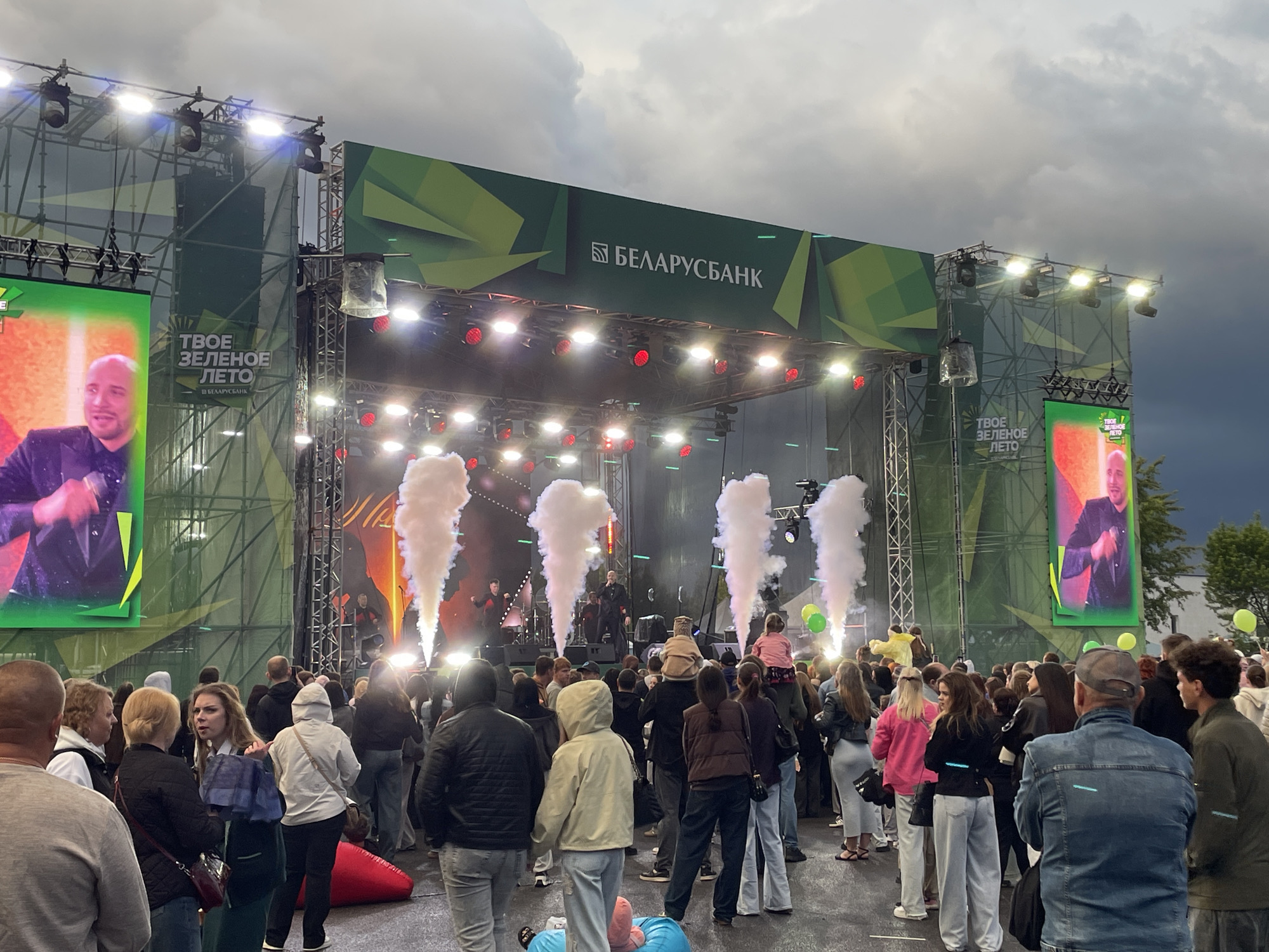 The image depicts an outdoor concert event, likely a festival, taking place in the city of Brest, Belarus. A large stage is set up with a prominent green banner overhead featuring the logo and name of "БЕЛАРУСБАНК" (Belarusbank) in white lettering, along with decorative green abstract shapes. Two large LED screens flank the stage. The left screen displays a live feed of a performer wearing a black jacket, while the right screen shows a similar performer.
On stage, several performers are visible, illuminated by stage lights and accompanied by plumes of white smoke emanating from the stage floor. The crowd is gathered in front of the stage, observing the performance. The attendees are diverse in age and attire, with many dressed casually. Some individuals are holding balloons, including bright green ones visible towards the right side of the frame. The overall atmosphere suggests a lively and engaging event. The sky is overcast, hinting at potentially damp weather. The background shows supporting structures for the stage and lighting equipment, with some greenery and trees visible on the far right. The ground surface appears to be asphalt or a similar paved material.
