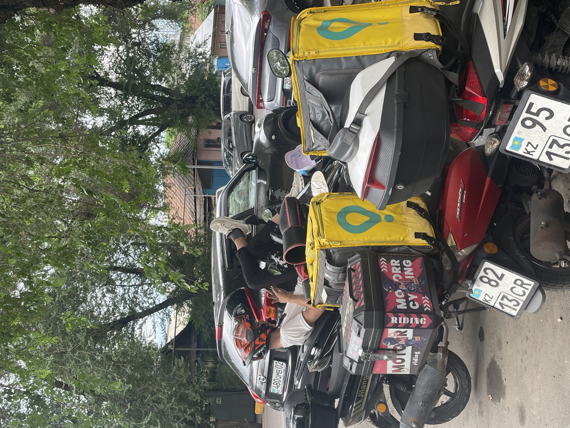 The image depicts a street scene in Almaty, Kazakhstan, likely on a day with overcast skies, as indicated by the diffused light and the lush green foliage of trees overhead. In the foreground, a person is seated on a black scooter, wearing a white t-shirt, black pants, and a distinctive orange and black motorcycle helmet. Their left leg is propped up on the scooter's storage compartment, and they are holding a mobile phone in their right hand.
The scooter is equipped with two large, bright yellow delivery bags with a teal logo on them, a common sight for food or package delivery services. Next to this scooter, other scooters are also visible, one red and one silver, both with storage boxes and license plates. The license plates are consistent with those used in Kazakhstan, featuring the KZ designation. One plate reads "KZ 82 13 CR" and another reads "KZ 95 13".
Behind the scooters, several cars are parked or stopped. One car is a dark grey hatchback with a visible license plate "KZ 480 OMB 02". Another car, a silver sedan, is partially visible. In the background, residential buildings with tiled roofs are present, along with more trees. The overall scene suggests a moment of rest or waiting, possibly during a delivery route in a residential or mixed-use urban area of Almaty.