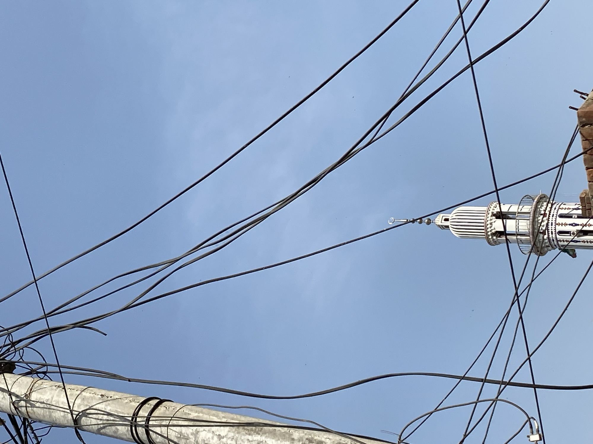 A low-angle, upward-looking photograph captures a dense network of black power lines and cables against a light blue, hazy sky. The sky, mostly clear but with a soft, diffused appearance, dominates the background.
To the right, a slender, white mosque minaret rises prominently. It is intricately designed with ornate patterns, possibly in contrasting dark colors, and features what appear to be tiered balconies. A distinct crescent moon finial crowns the very top of the minaret. Part of a brick structure, likely an adjacent building, is visible at the extreme top right corner, showing exposed brickwork and some protruding metal elements.
In the bottom left of the frame, a section of a weathered concrete pole or pillar is visible. Numerous black cables are tightly wrapped around this pole and extend from it, crisscrossing diagonally across the sky. The wires vary in thickness and create a complex, somewhat tangled pattern.
The scene depicts a blend of religious architecture and utility infrastructure, characteristic of urban and semi-urban environments in locations like Chak 166 GB, Pakistan. There are no people visible, nor is there any discernible text. The diffused light indicates daytime, possibly under an overcast or hazy sky.