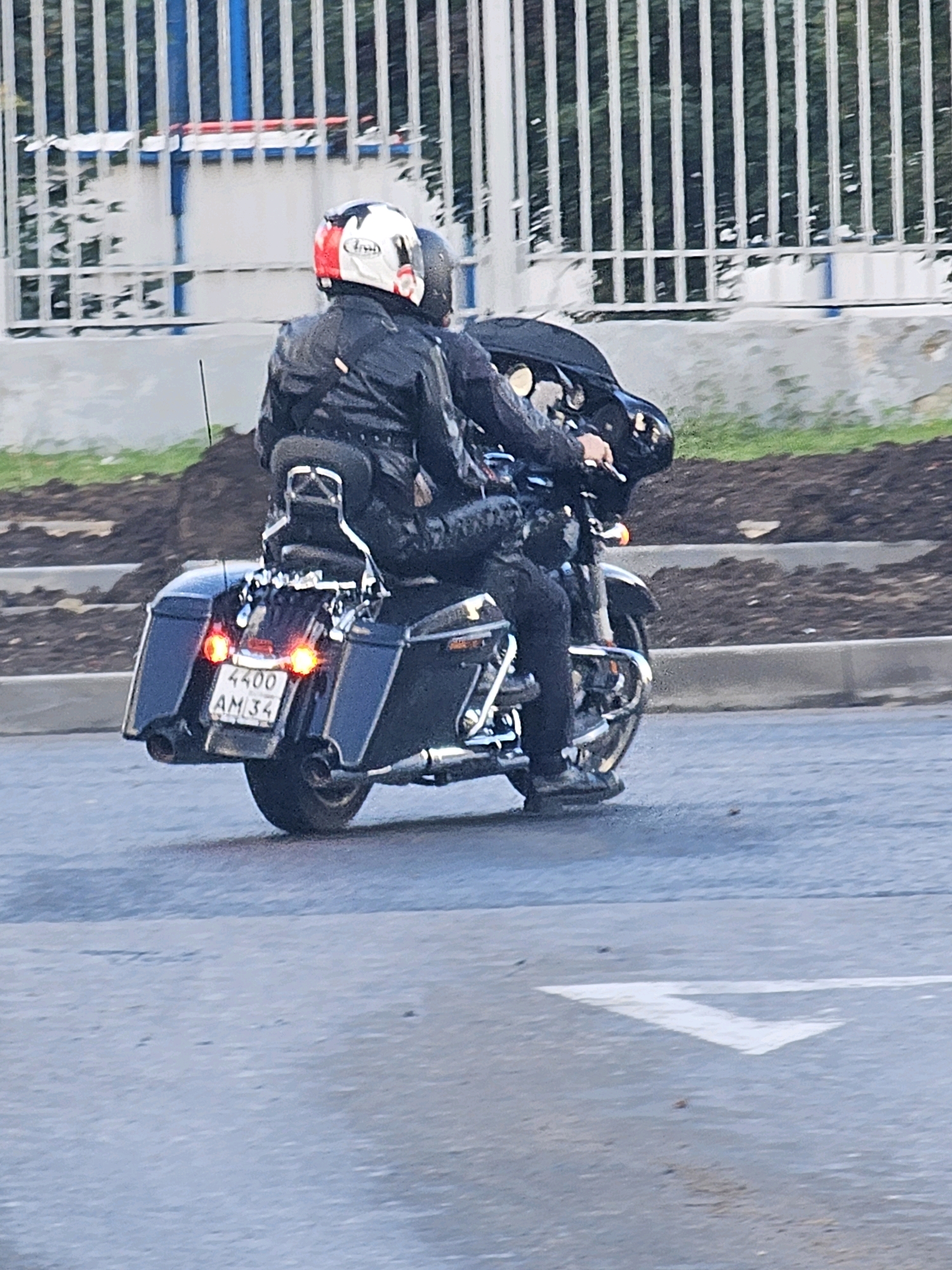 A motorcycle carrying two people is captured in motion on an asphalt road. The motorcycle is black with chrome accents, and has saddlebags on the rear. The license plate is visible, reading "4400 AM 34". The rider in front wears a black leather jacket and helmet with red, white, and black detailing. The passenger behind also wears black clothing and a dark helmet. The background features a white and blue fence and some greenery. The image appears to be taken in the city of Khimki, Russia, based on the license plate format.