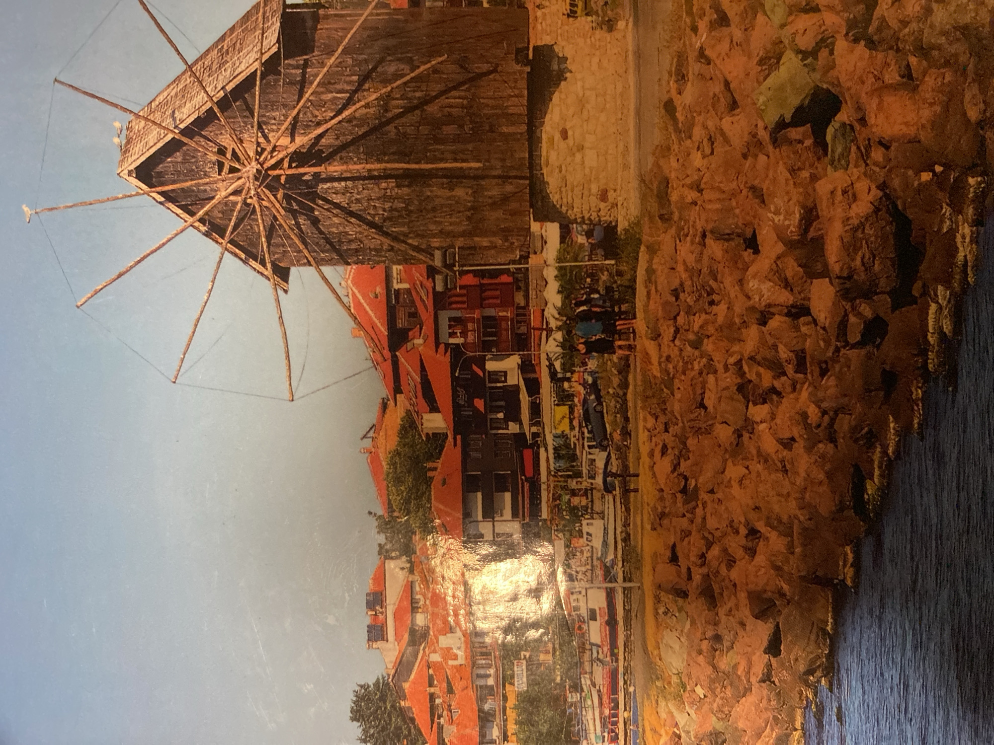 The image depicts a landscape with a traditional windmill on the left side, positioned above buildings with red-tiled roofs. The buildings are situated along what appears to be a street or promenade with some activity. On the right, there is a rocky or stone embankment leading to a body of water. The sky is visible in the upper-left corner, suggesting it is daytime. The specific location is not evident from the image alone.
