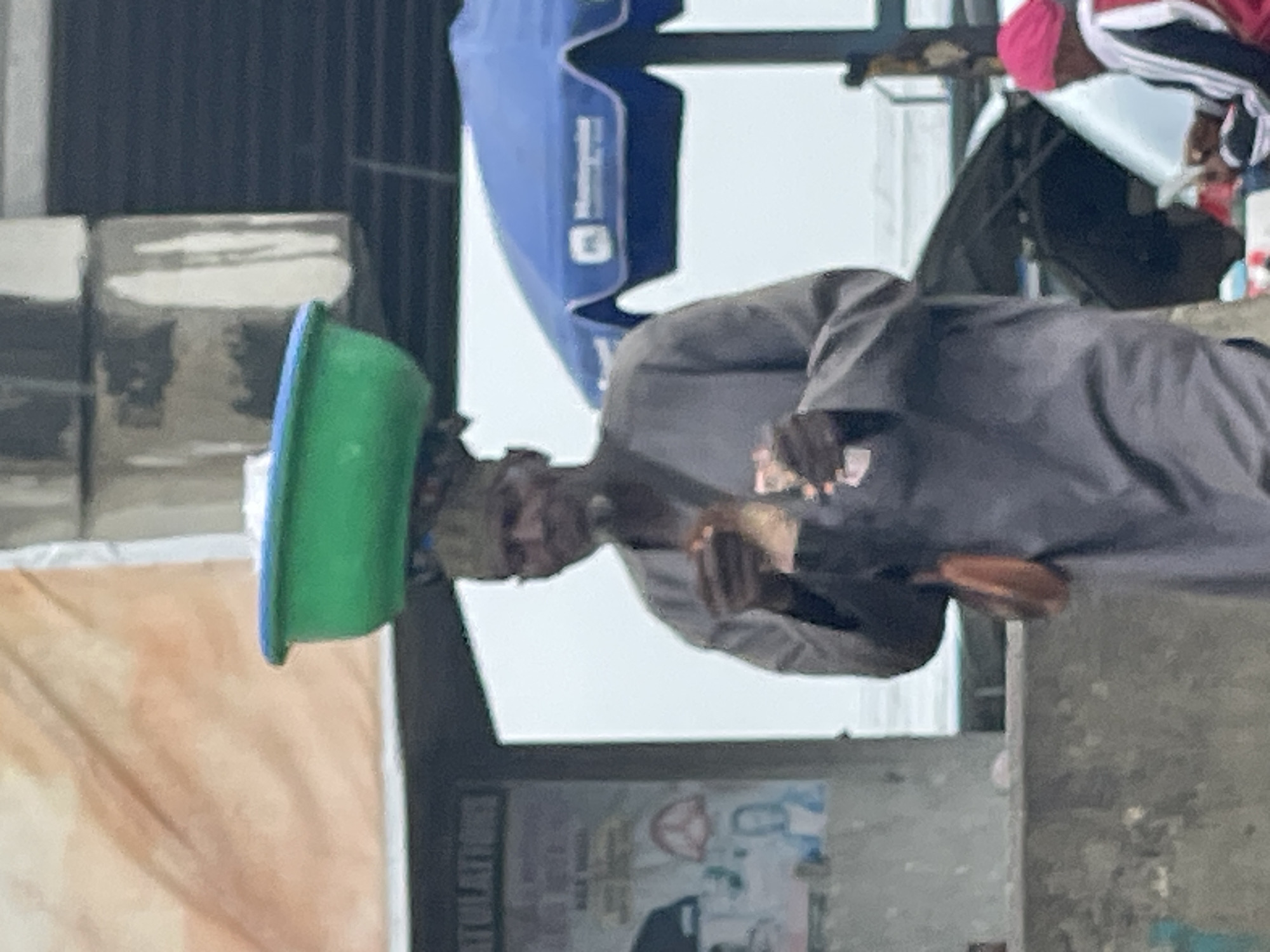 A man is seen in an outdoor urban setting, balancing a green tub on his head. He is wearing a dark-colored, long-sleeved garment. The man is standing near a concrete structure with what appears to be posters affixed to it. An open blue umbrella is partially visible in the background. Another person wearing a pink head covering and a jacket with black and white stripes is also visible. The scene is located in Ado, Nigeria.