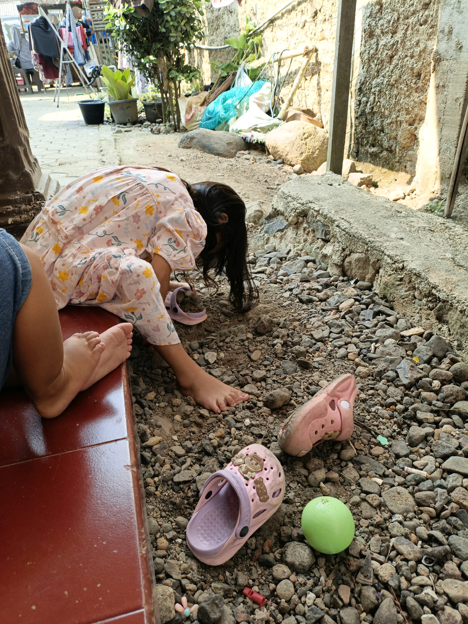 A young girl in a floral-patterned dress is hunched over on a pebbly ground. Her dark hair cascades over her face as she appears to be playing with the ground. To her left, another child's legs are visible, wearing blue jeans and sitting on a reddish-brown tiled surface. Scattered around the girl are two pink children's clogs, a bright green ball, and various small rocks. In the background, laundry hangs on a rack and various potted plants are visible, suggesting a residential setting. The scene is outdoors, with sunlight casting shadows. The location is identified as Bunut, Indonesia.