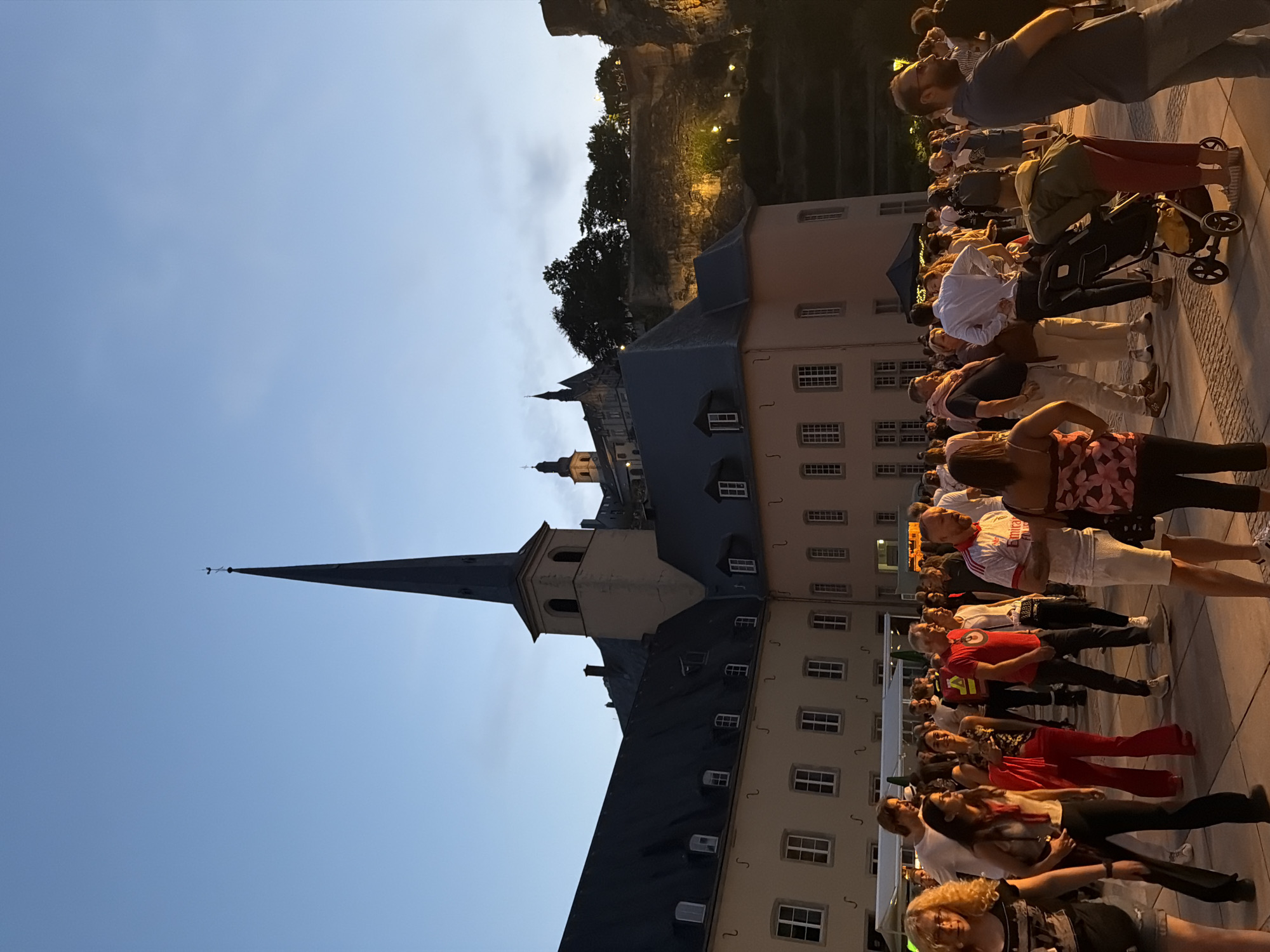 The image depicts a large outdoor gathering in front of a historic building in Luxembourg City. The architecture suggests a significant cultural or religious site, possibly a former monastery or church complex, with a prominent steeple rising against the twilight sky. A crowd of people, diverse in age and attire, are milling about in the foreground and midground. Some are casually conversing, others appear to be observing the scene, and a few are in motion. Among the attendees, a person is pushing a stroller, indicating families are present. The setting appears to be a public square or courtyard, paved with stone. The overall ambiance is that of a communal event or festival, illuminated by the fading daylight and artificial lights on the distant cliffside structures.