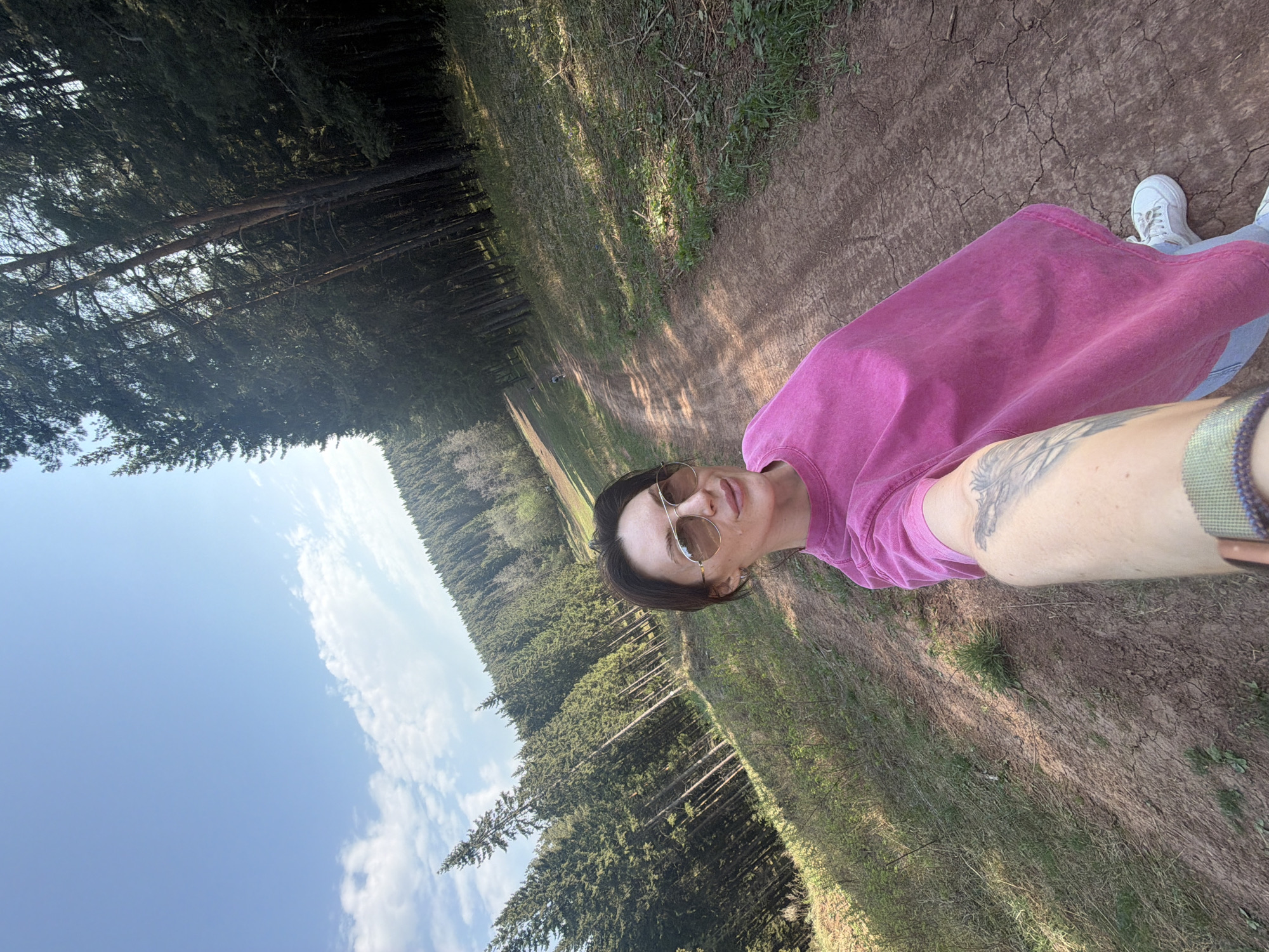 A woman is posing for a selfie on a dirt trail bordered by a dense forest. The trail is dry and cracked, with patches of grass. The forest consists mainly of evergreen trees. The sky is visible with some light clouds. The woman is wearing sunglasses and a pink shirt. She has a tattoo on her left forearm. She is wearing white sneakers and light blue jeans. The location is likely in a forested area on the outskirts of Perm, Russia, but this cannot be definitively confirmed.