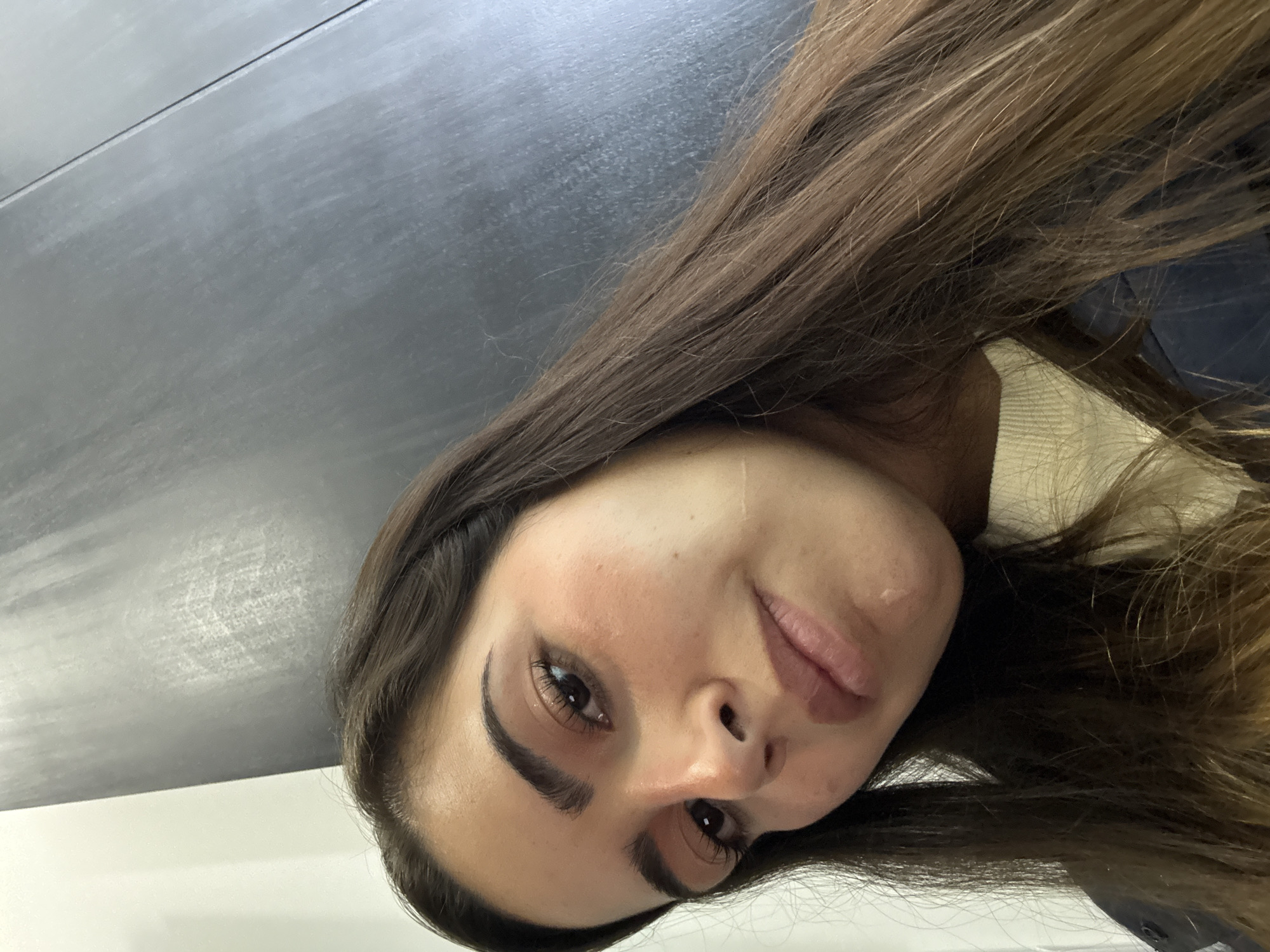 The image is a close-up, tilted selfie of a light-skinned woman with long brown hair. The background consists of a dark, reflective surface and a white wall. No details suggest this image was taken in San Bernardino Tlaxcalancingo, Mexico.