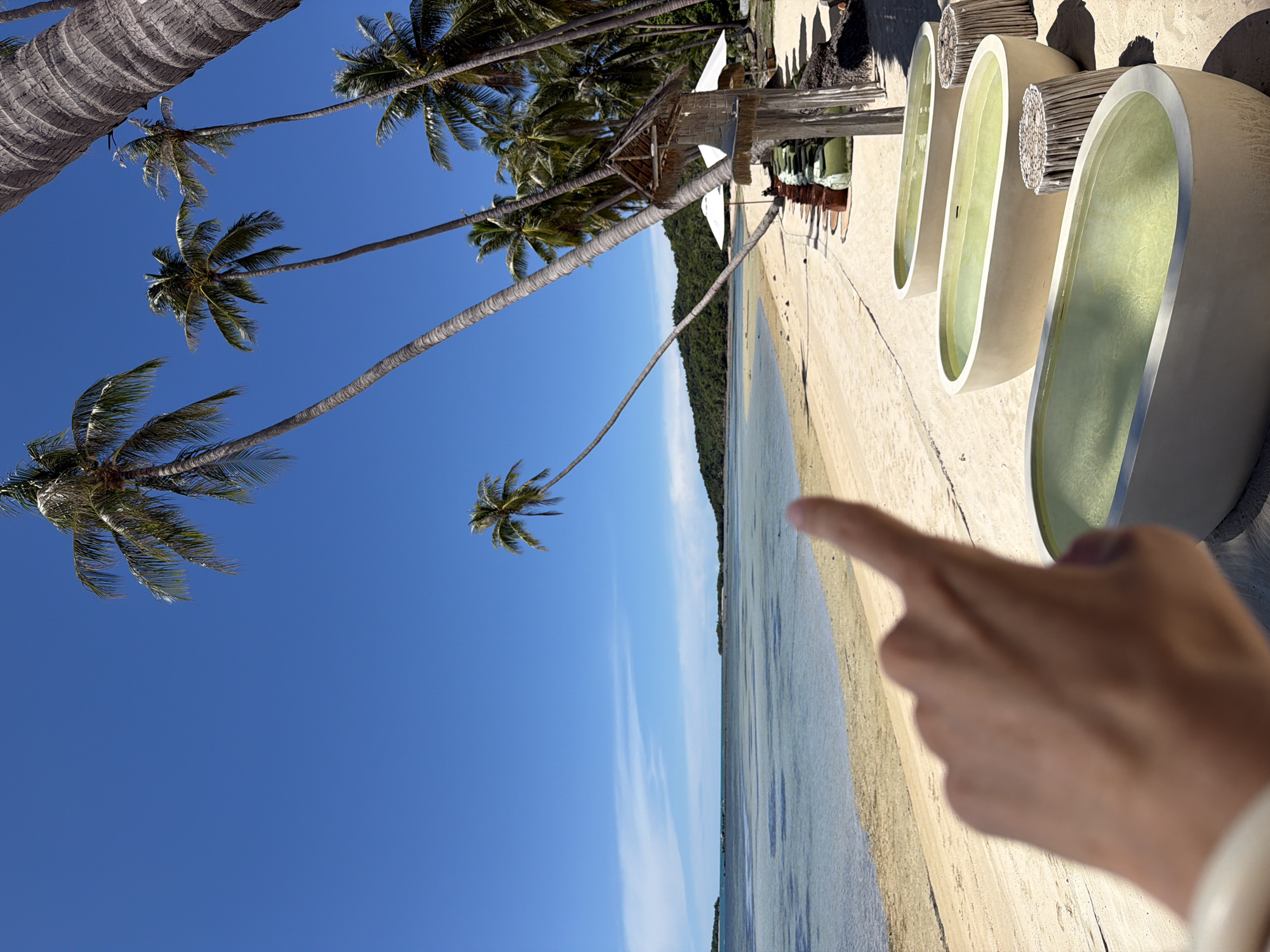 The image depicts a serene beach scene in Baan Thong Krut, Thailand. The foreground is dominated by a person's hand, with a finger pointing towards the ocean, suggesting an interaction with the environment or a directorial gesture. The scene transitions to a tranquil beach with calm, shallow waters. To the right, a row of four oval-shaped, light-colored bathtubs filled with a pale green liquid are visible, set against a sandy area and surrounded by wooden structures and bundles of reeds. These bathtubs appear to be part of a resort or spa, offering a unique relaxation experience by the sea.
Tall palm trees, with their characteristic trunks and fronds, are scattered throughout the image, framing the view and emphasizing the tropical setting. The sky is a clear, vibrant blue, indicative of a sunny day. The water is a soft turquoise, reflecting the bright sky and creating a sense of peace and tranquility. In the distance, a lush green hillside or island is visible, adding depth to the landscape. The overall atmosphere conveyed is one of relaxation, natural beauty, and a luxurious tropical getaway. No visible text is present in the image.