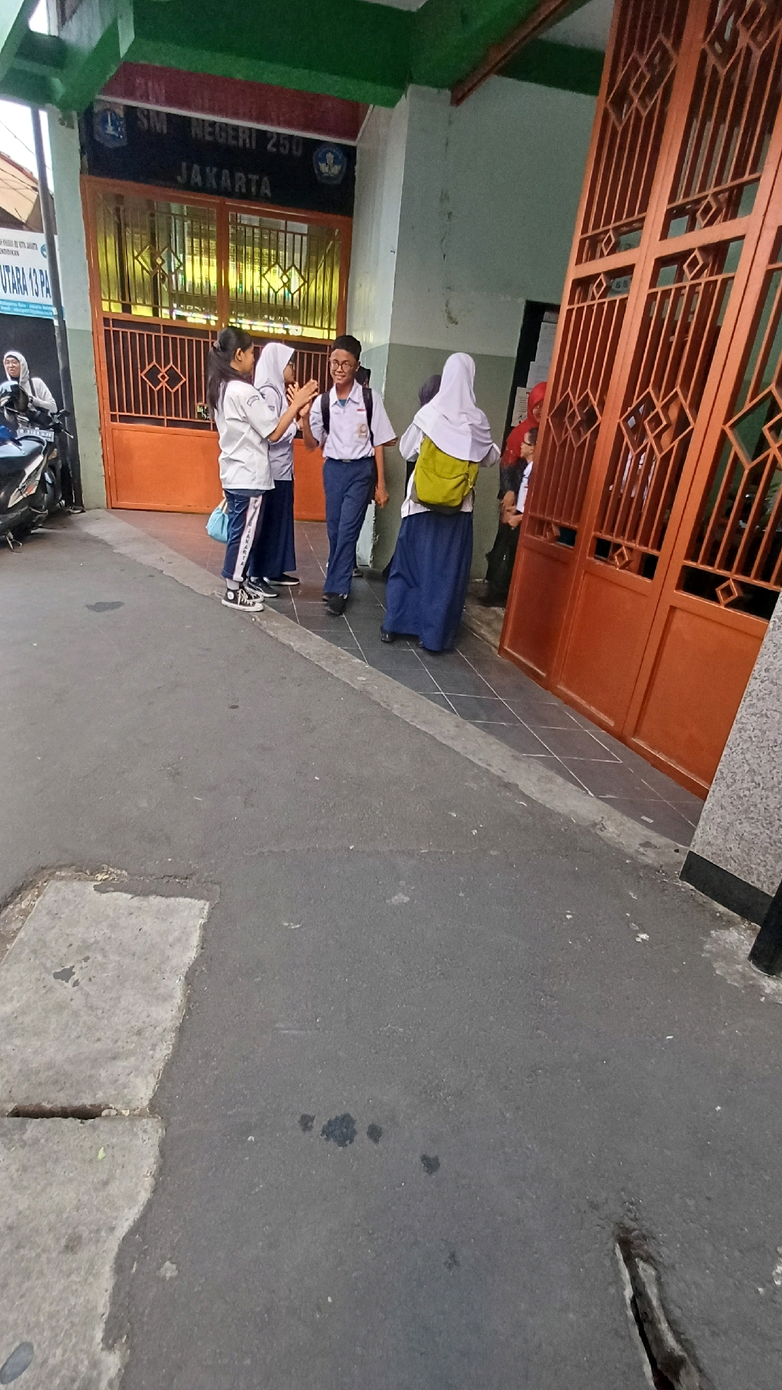The image captures a scene outside a school building with orange gates and a green overhang. At the entrance, the text "SM NEGERI 250 JAKARTA" is visible. Four students, dressed in school uniforms consisting of white shirts and blue skirts or pants, are interacting near the gate. Two students are facing each other, appearing to be talking or greeting, while another student in a white hijab and a yellow backpack stands nearby, facing away from the camera. A fourth student in a white hijab and blue skirt is also visible. To the left of the school entrance, a motorcycle is parked on the sidewalk. The ground in front of the school is paved with asphalt and features a concrete slab with a crack. The overall setting suggests an outdoor area in front of an educational institution in Jakarta, Indonesia.