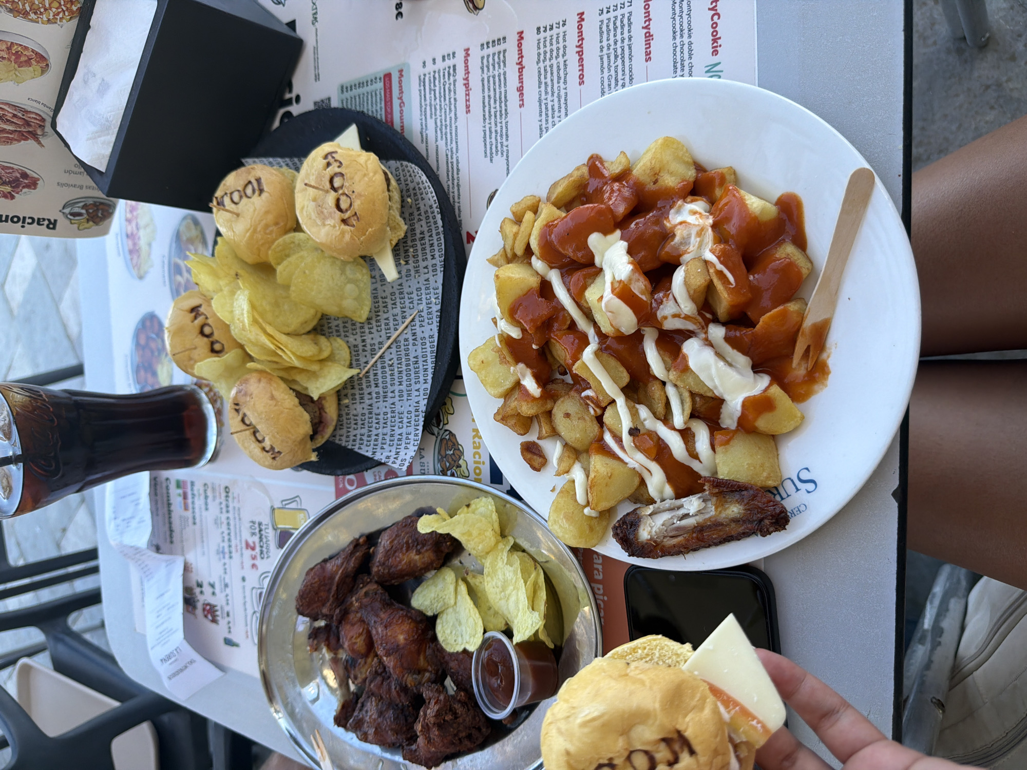 This image shows a table covered with menus and food items at what appears to be a restaurant in Alicante, Spain. The table has several dishes including:
- A black tray of mini burgers with "KOOL" branded on the buns, accompanied by potato chips.
- A white plate of patatas bravas, covered in red and white sauce, with a piece of what appears to be chicken.
- A steel bowl containing a collection of fried chicken wings, with potato chips, and a small cup of sauce.
- A hand holding a mini burger with cheese.
There is a glass of dark liquid, possibly cola, on the table along with receipts or order sheets.
The location is inferred due to text references in menus in the image.