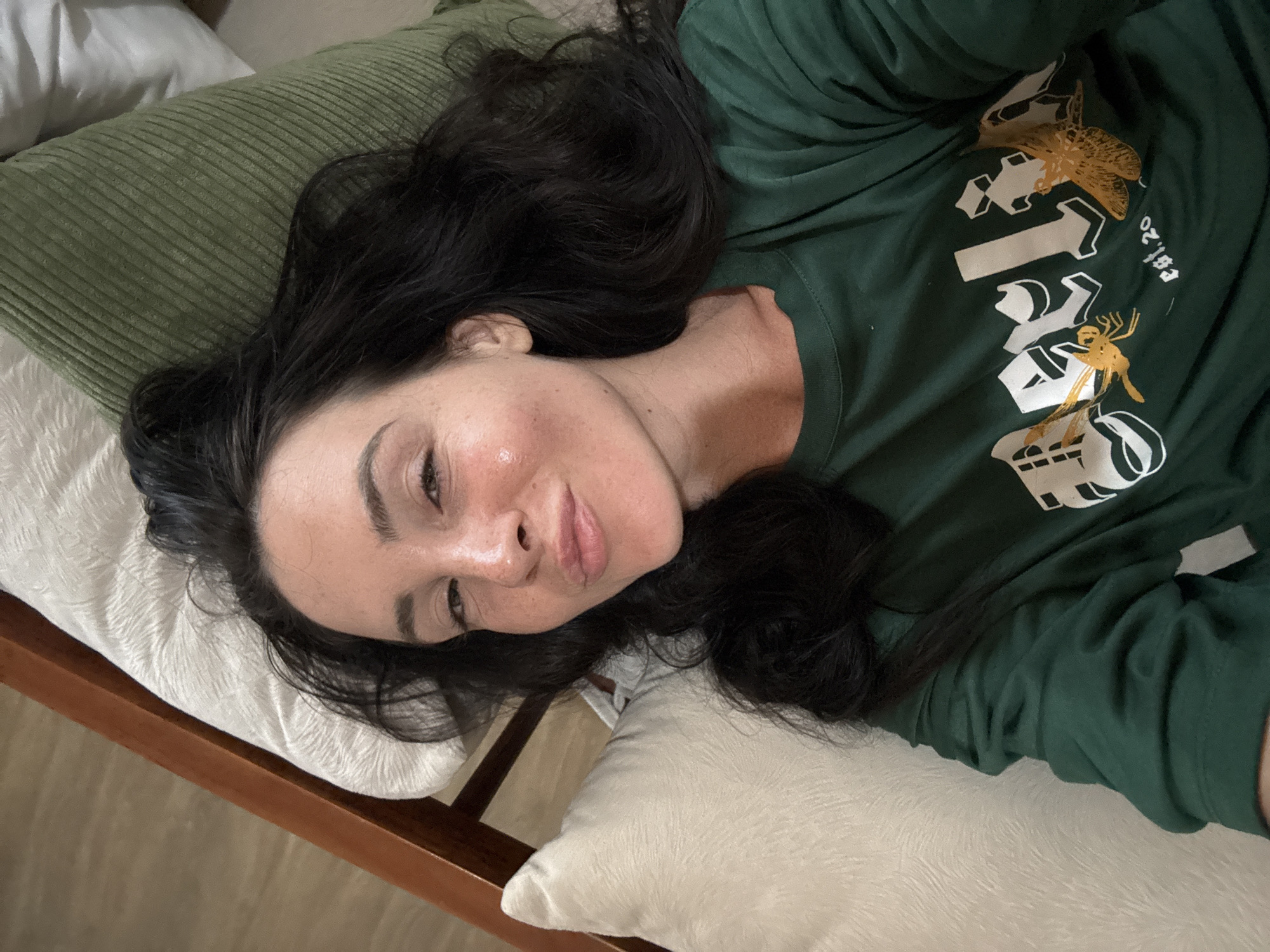 A woman with long, dark hair is lying down, propped up by pillows. She is wearing a dark green t-shirt with white and gold graphics. Her head is tilted back slightly and she is looking towards the camera with a slight smile. The background consists of pillows and a wooden headboard. The image appears to be taken indoors in a bedroom.