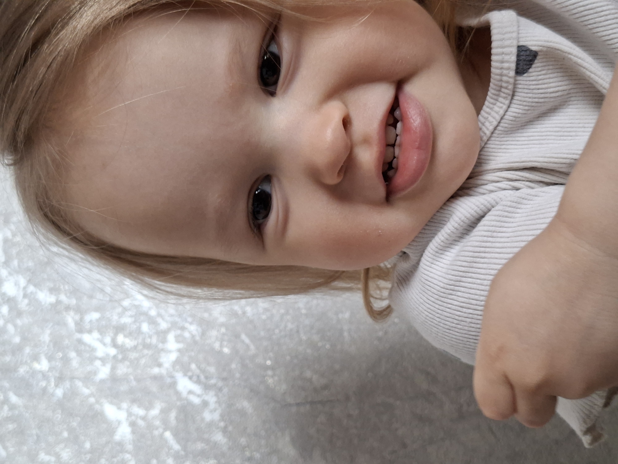 A close-up image depicts a baby with blonde hair and a wide, toothy smile. The baby's face is slightly tilted to the side, with large, dark eyes and rosy cheeks. The baby is wearing a striped white shirt with a small heart near the collar. The background is a textured, light-colored surface. The location is Unknown, Unknown.