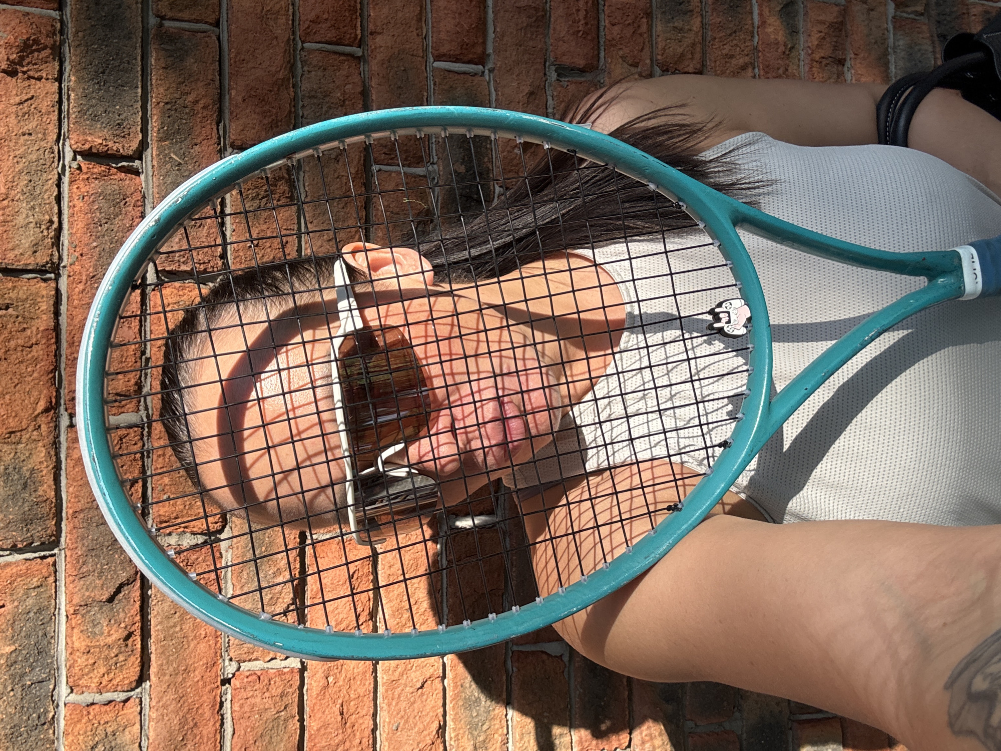 A woman is lying on her side against a brick wall. She is wearing a white tank top. A blue tennis racket is held in front of her face. The location is Chaikovsky, Russia.