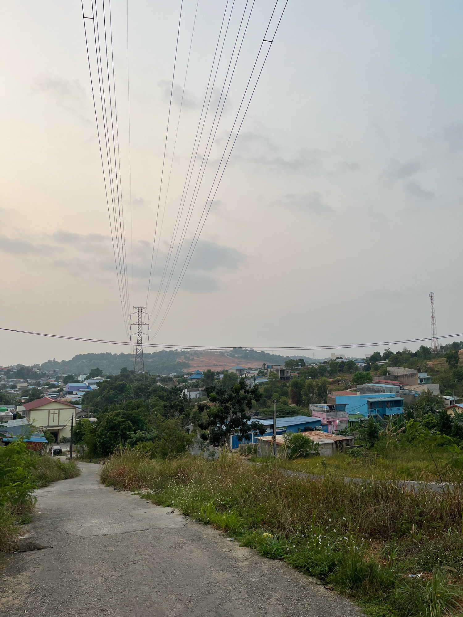 A rural road leads uphill towards a cluster of houses and a large power line tower in Batam, Indonesia. The sky is overcast and hazy, obscuring the horizon. The road itself is paved but cracked and appears to be in disrepair, with overgrown grass and weeds lining its sides. In the mid-ground, a collection of colorful houses, mostly single-story with brightly colored roofs, are nestled amongst lush green trees. The prominent power line tower stands tall in the distance, with multiple cables fanning out and disappearing into the hazy sky. A telecommunications tower is visible on the right side of the image. The overall scene suggests a developing area on the outskirts of a city, characterized by both infrastructure and the natural landscape.