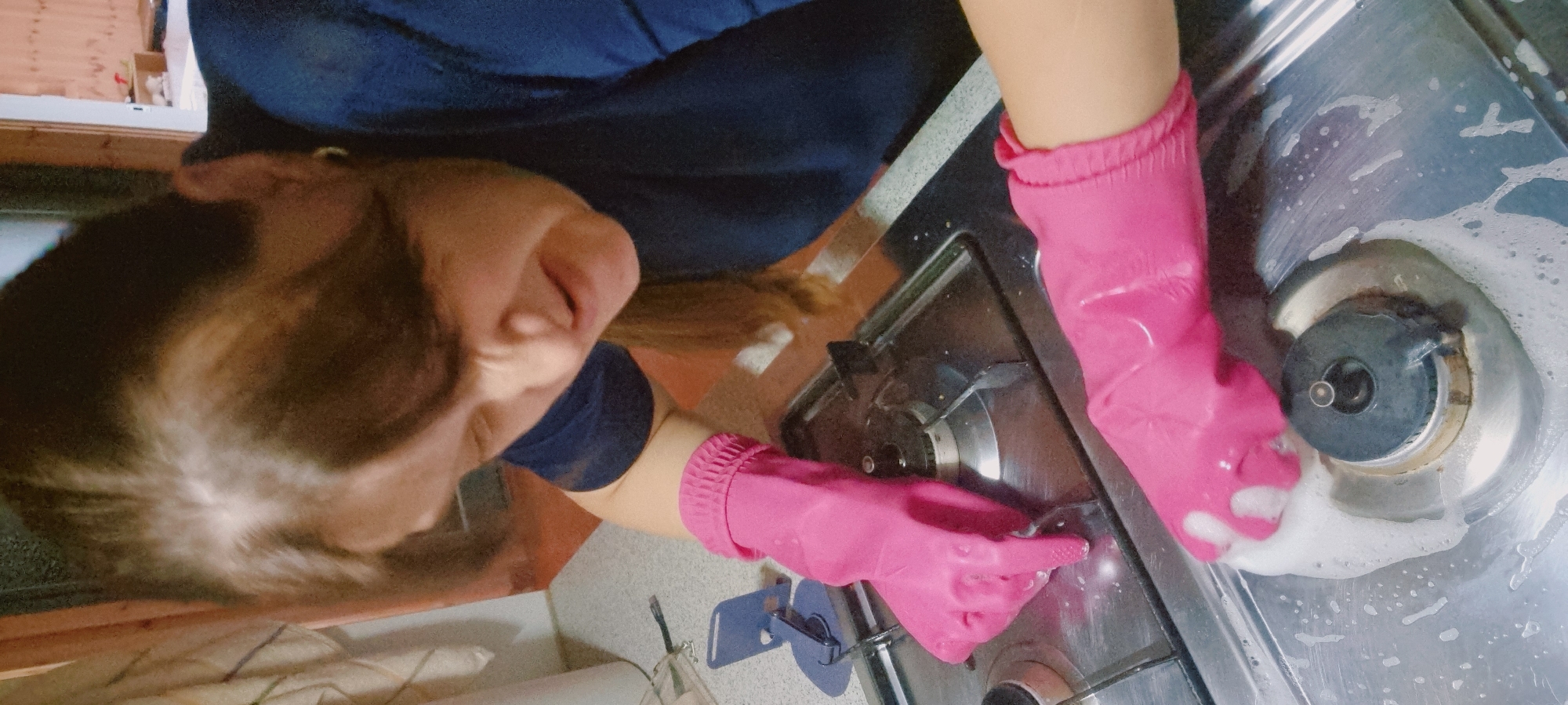 A person wearing pink gloves is cleaning a stovetop with soapy water. The person has dark hair and is wearing a dark blue shirt. The location is likely in Dunnae-myeon, South Korea. The stovetop is silver and has multiple burners. There is soap suds on the stovetop.