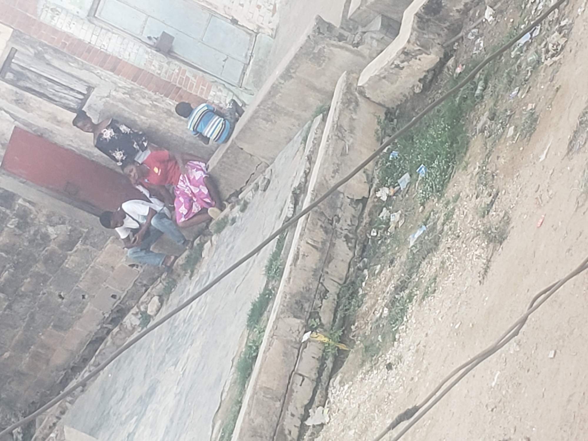 A ground-level, slightly out-of-focus shot shows a group of people sitting outside a building in Alalubosa, Nigeria. A man in a white shirt and jeans is seated on a step. Next to him is a person in a red shirt, followed by another in a pink patterned dress. Further along, a woman in a black floral top stands. Lastly, there's a person in a blue striped shirt seated next to the building. The building itself has boarded-up windows, a red door, and aged brickwork. Adjacent to the building is a patch of dirt with sparse grass and litter. Two power lines run across the foreground. A yellow bird is near the curb.