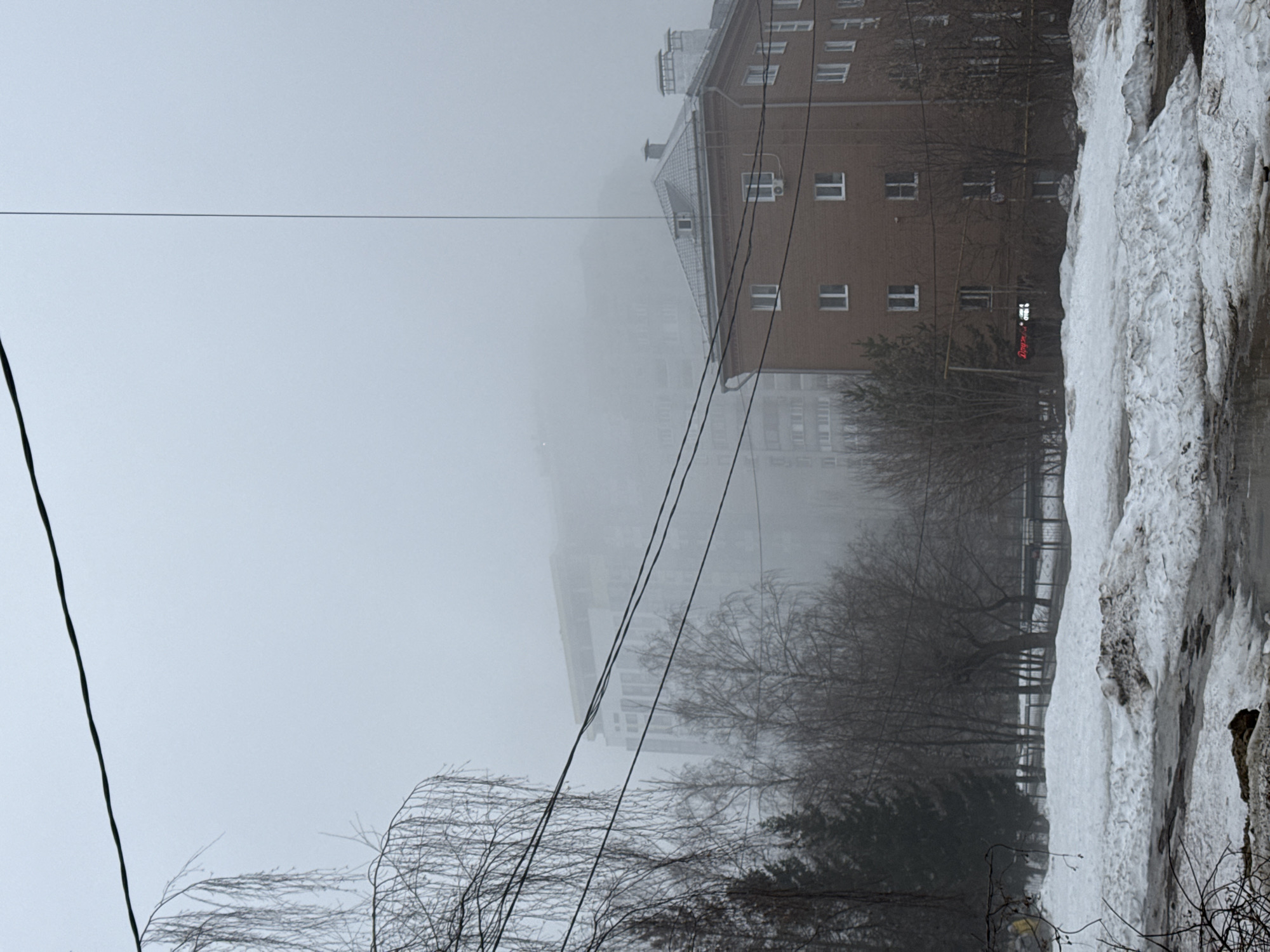 The image depicts a foggy street scene in Kazan, Russia, likely during winter. A multi-story brick building with white-framed windows is visible, partially obscured by the fog. Power lines traverse the scene. Trees with bare branches are present. A significant amount of snow and ice lines the right side of the image. A building in the fog has a bright red sign, possibly a store of some kind.