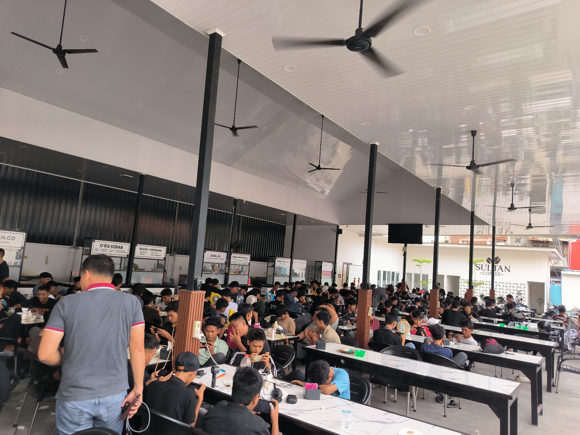 The image shows a bustling scene in a large, open-air restaurant, likely in Lhokseumawe, Indonesia. Numerous patrons, predominantly young men, are seated at long tables, many engrossed in their mobile phones, suggesting a digital engagement. The setting features high ceilings with multiple ceiling fans. Signage for food vendors, including "D'RIZ KEBAB" and "BAKSO TARIWANG", along with "SULTAN -COFFEE-" indicates a food court or collective dining space. A person in a grey shirt and jeans is walking through the space. Tables are set with water glasses and charging stations.