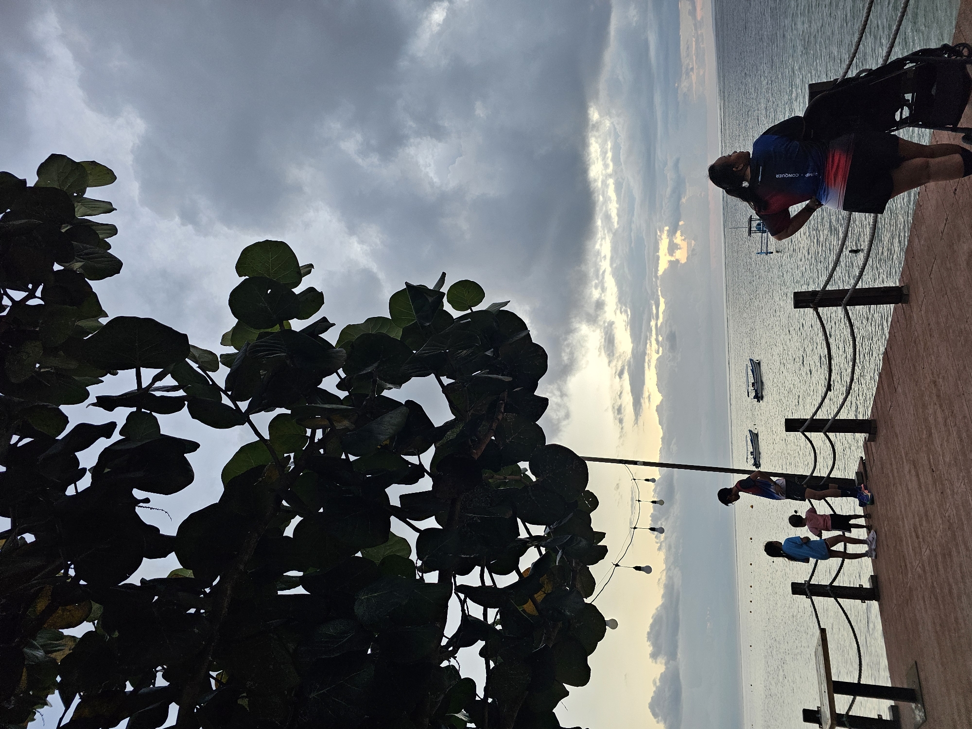 The image depicts a family on a wooden pier in Denpasar, Indonesia, by the coast. In the foreground, a woman stands with her hands on her hips, looking out at the ocean, with a stroller beside her. Further down the pier, a man stands with two children, a girl in a blue shirt and shorts and a younger child in a pink shirt and shorts. A table is visible on the left side of the pier. String lights are hung above the pier, and boats are scattered on the water. The sky is cloudy, with a hint of sunset colors. The overall scene suggests a casual evening outing by the sea.