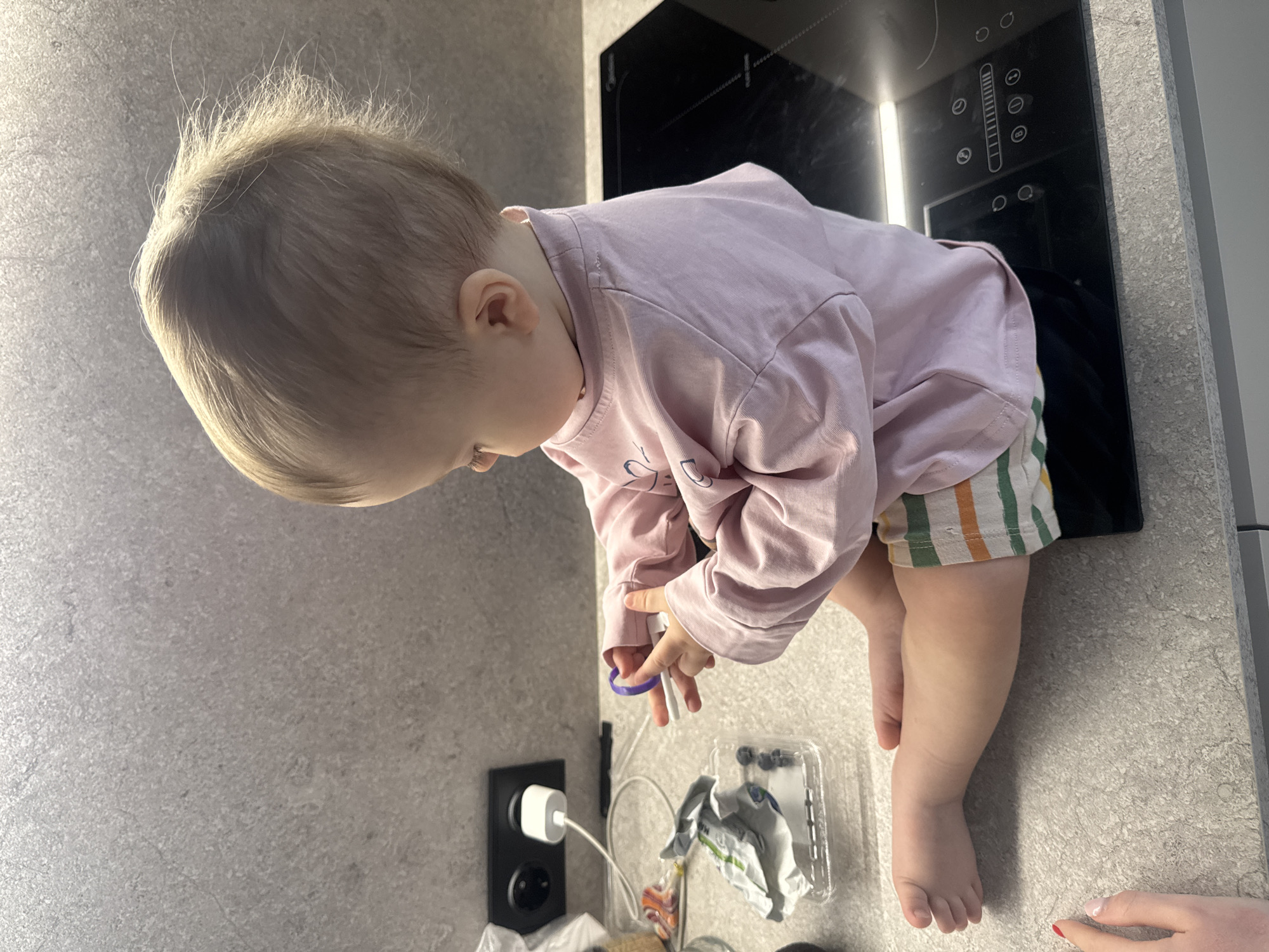 A curious baby with light blonde, wispy hair is captured leaning over a speckled grey kitchen countertop, intently focused on objects in their hands. The baby is wearing a light pink long-sleeved shirt and striped shorts with green, orange, and white bands. In their hands, they hold a small purple ring-like item and a white stick-like object.
The background features a modern kitchen setup with a black induction cooktop, which displays various illuminated touch controls, including a vertical slider, circles, and symbols. Faint text "Midea" and "PowerBoost" are visible on the cooktop's surface. To the left of the baby, a black wall outlet with a white charger and accompanying cables is visible. Nearby, a clear plastic container, possibly containing blueberries, and a crumpled white wrapper are placed on the counter.
An adult's hand with red nail polish on a fingertip is partially visible in the bottom right corner, suggesting supervision or interaction. The scene is brightly lit, likely by natural light, casting soft shadows. The overall impression is a moment of childish exploration within a home kitchen setting.