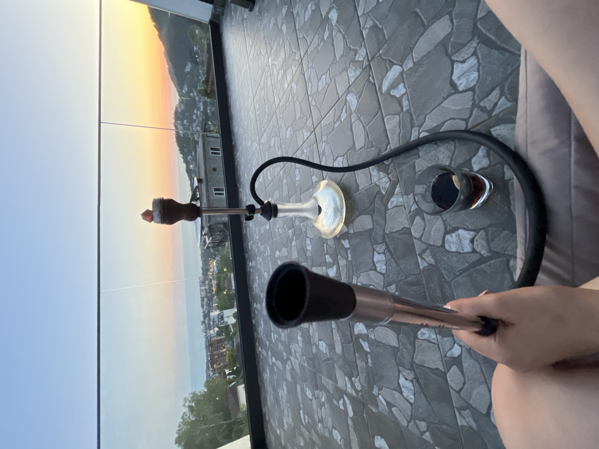 The image captures an elevated outdoor perspective at sunset or sunrise in Baranovka, Russia. The foreground shows a seated individual's lower legs and a right hand holding a black and metallic silver hookah mouthpiece. A black hookah hose extends from the mouthpiece across the grey, textured tiled floor. Adjacent to the hose, a clear glass tumbler containing a dark liquid and a lemon slice rests on the tiles.
In the midground, a modern hookah stands upright on the tiled surface. It features a clear glass base, a slender metallic stem, and a dark ceramic bowl atop the stem. The black hose connects to the hookah's base.
Beyond the tiled platform, a glass railing with metallic supports provides an unobstructed view of a sprawling cityscape nestled against a backdrop of undulating, tree-covered hills and mountains. A large body of water, likely the Black Sea, is visible beyond the city and hills. The sky displays a gradient of colors, transitioning from light blue overhead to warm orange and yellow hues at the horizon, indicative of either sunset or sunrise. Buildings of various sizes are dispersed throughout the urban landscape.