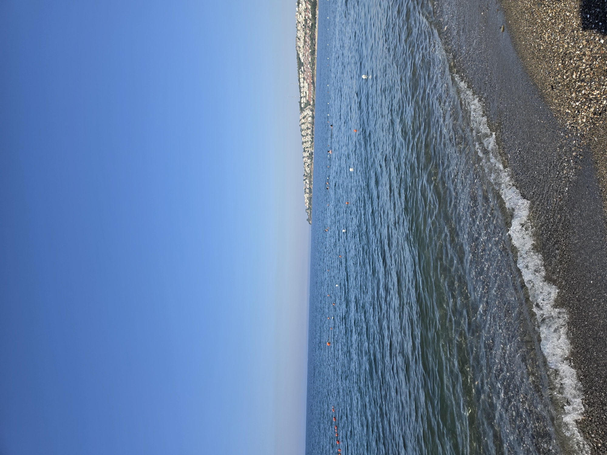 The image depicts a serene beach scene on a clear, sunny day. The foreground shows a pebble beach with a gentle wave lapping at the shore. The water is a deep blue, with some areas of lighter, clearer water near the beach, revealing the pebbles beneath. Scattered in the water are buoys, demarcating a swimming area. In the distance, a coastline is visible, lined with white buildings that ascend a gentle slope, characteristic of Seferihisar, Turkey. Some people are visible in the water, appearing to be swimming or enjoying the sea. The sky is a vast expanse of clear, bright blue, indicating fair weather. The overall impression is one of a peaceful, coastal resort town.