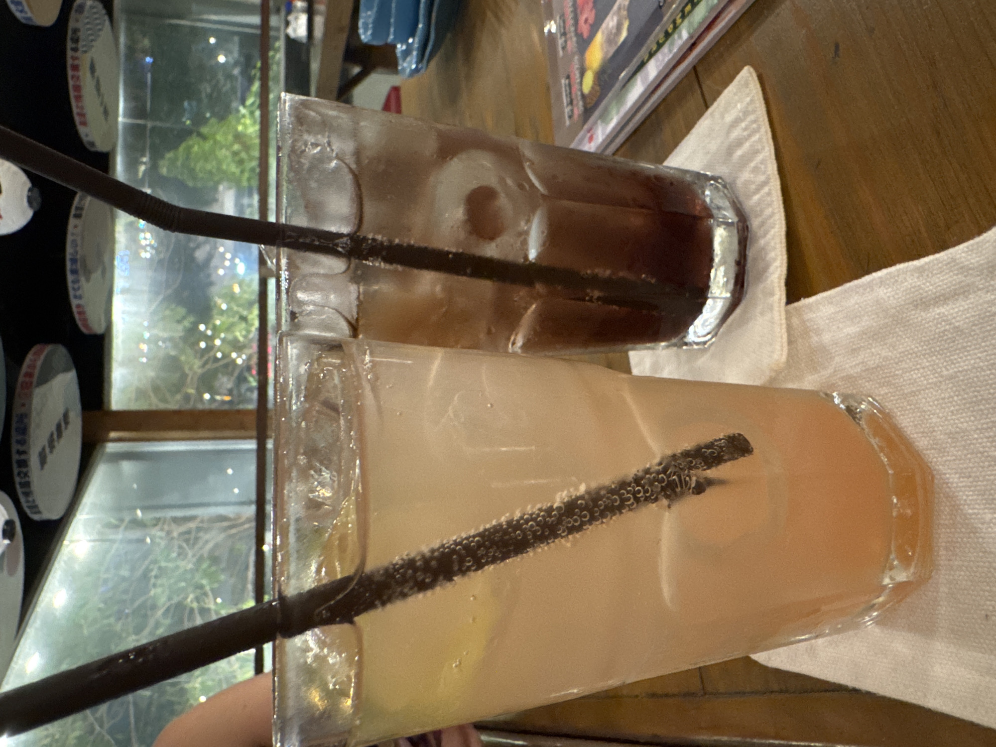 On a wooden table sits two tall, clear, multifaceted glasses. The glass closer to the viewer contains a light orange liquid with ice and a dark brown straw. The glass behind it contains a dark brown liquid with ice and a dark brown straw. Both glasses are on white linen napkins. To the left, a window shows the outside with greenery and blurred city lights. Based on the urban setting and the style of the dishes visible through the window, it is likely located in Bangkok, Thailand. The atmosphere suggests an indoor setting, such as a restaurant or bar.
