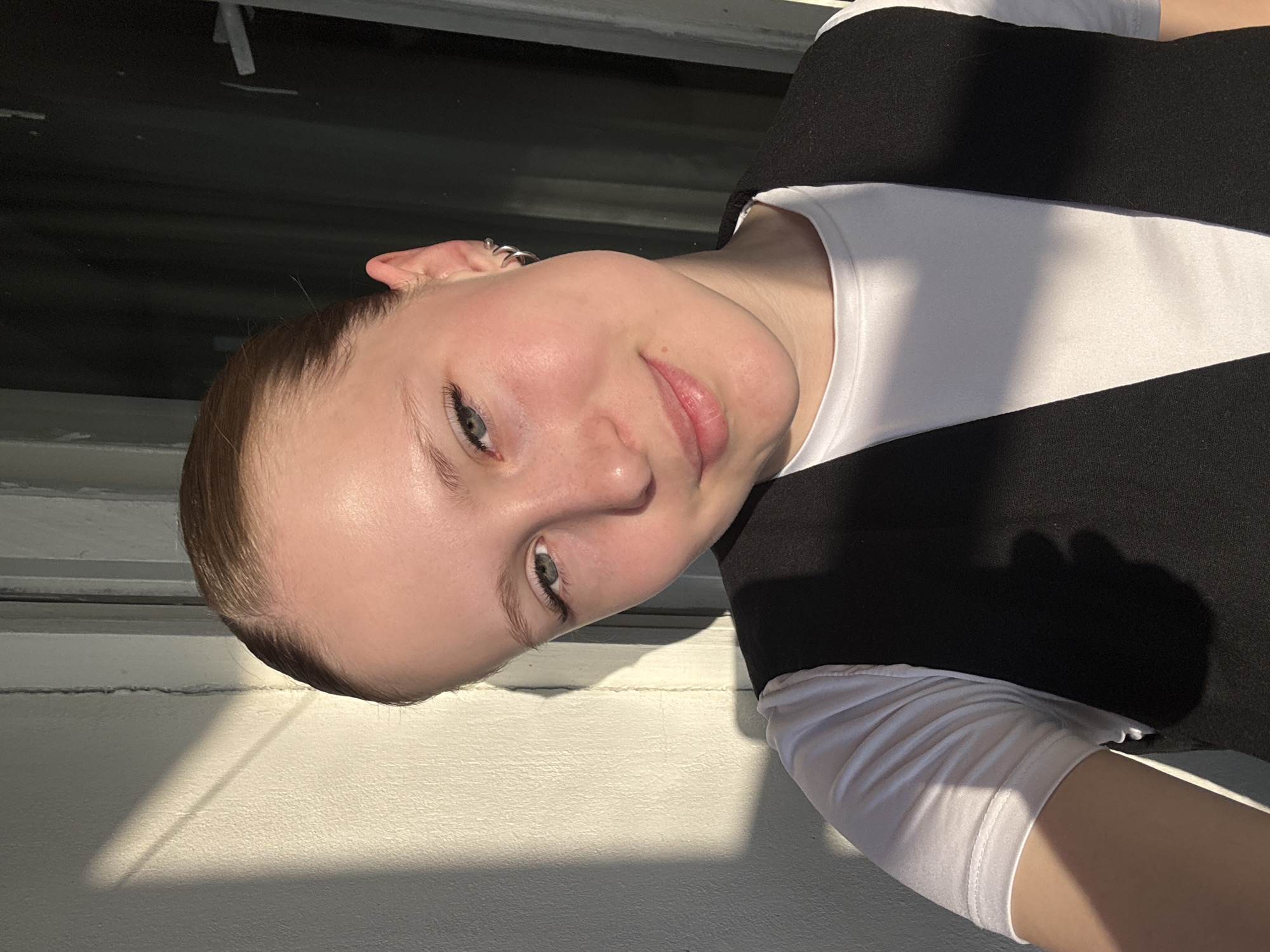 The image is an indoor shot of a young woman. She has light skin, light-colored eyes, and her hair is pulled back. She is wearing a white shirt with a black vest. The lighting is bright, likely due to sunlight coming through a window. The background is out of focus, but appears to show a window frame and a white wall. There is nothing in the image to indicate its location in Chaikovsky, Russia. The scene is a simple portrait with a focus on the woman's face and upper body.