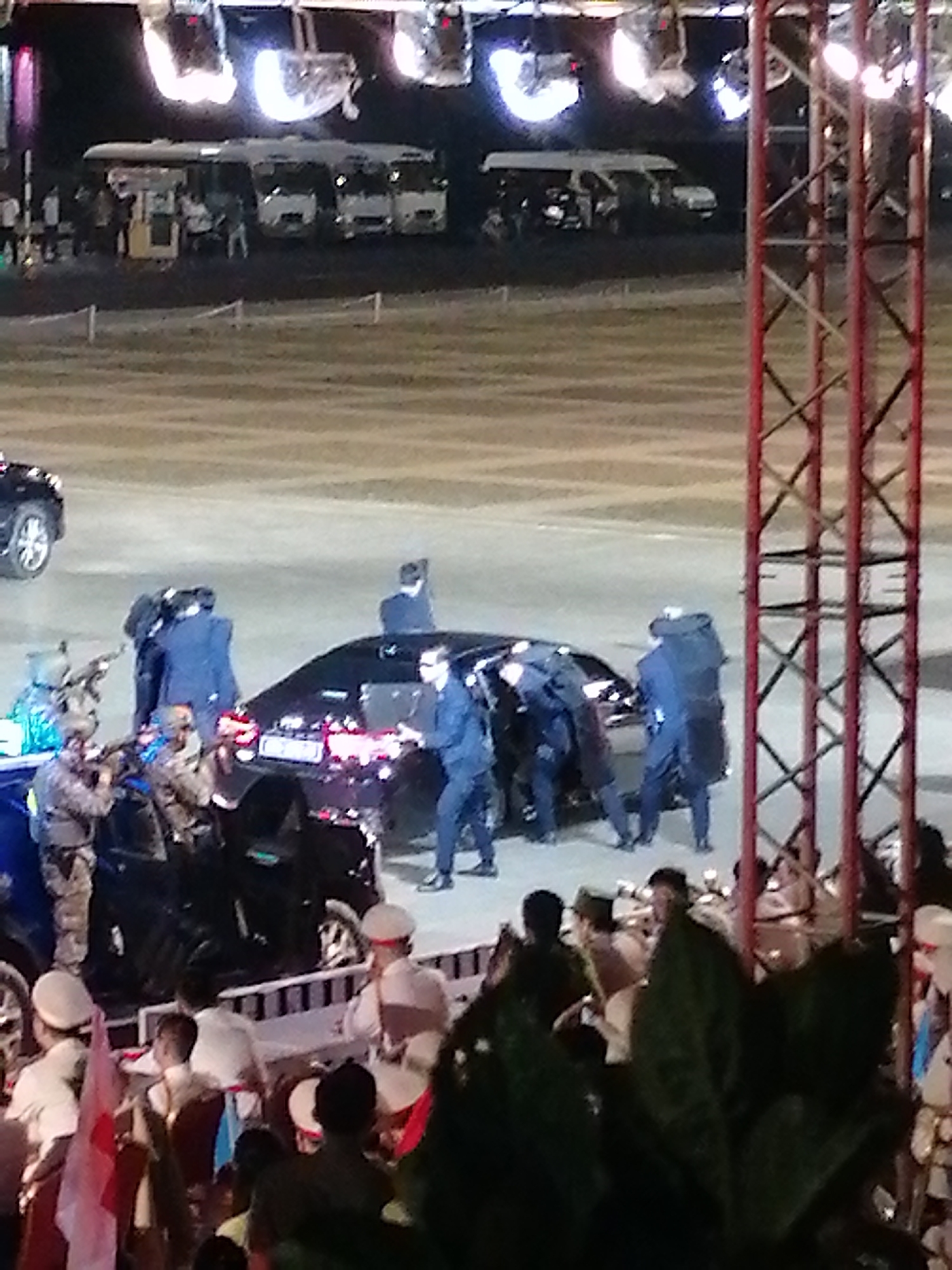 The image depicts a scene at night in Da Lat, Vietnam, likely at an outdoor event. The foreground shows a crowd of people, some wearing white hats and uniforms, others in casual attire. Flags are visible among the spectators, including a red and white flag, possibly the Vietnamese flag. In the midground, several individuals in dark suits, appearing to be security personnel, surround a black car. Some are positioned near the car's doors, and one person is leaning into the vehicle. Another person in a blue suit is standing near the car. In the background, a row of buses are parked, illuminated by overhead lighting. A large red metal structure, possibly a stage or scaffolding, is visible on the right side of the image. The overall atmosphere suggests a formal event or arrival, possibly involving dignitaries. The lighting indicates it is nighttime, and the weather appears to be clear. There are no discernible signs or text in the image.