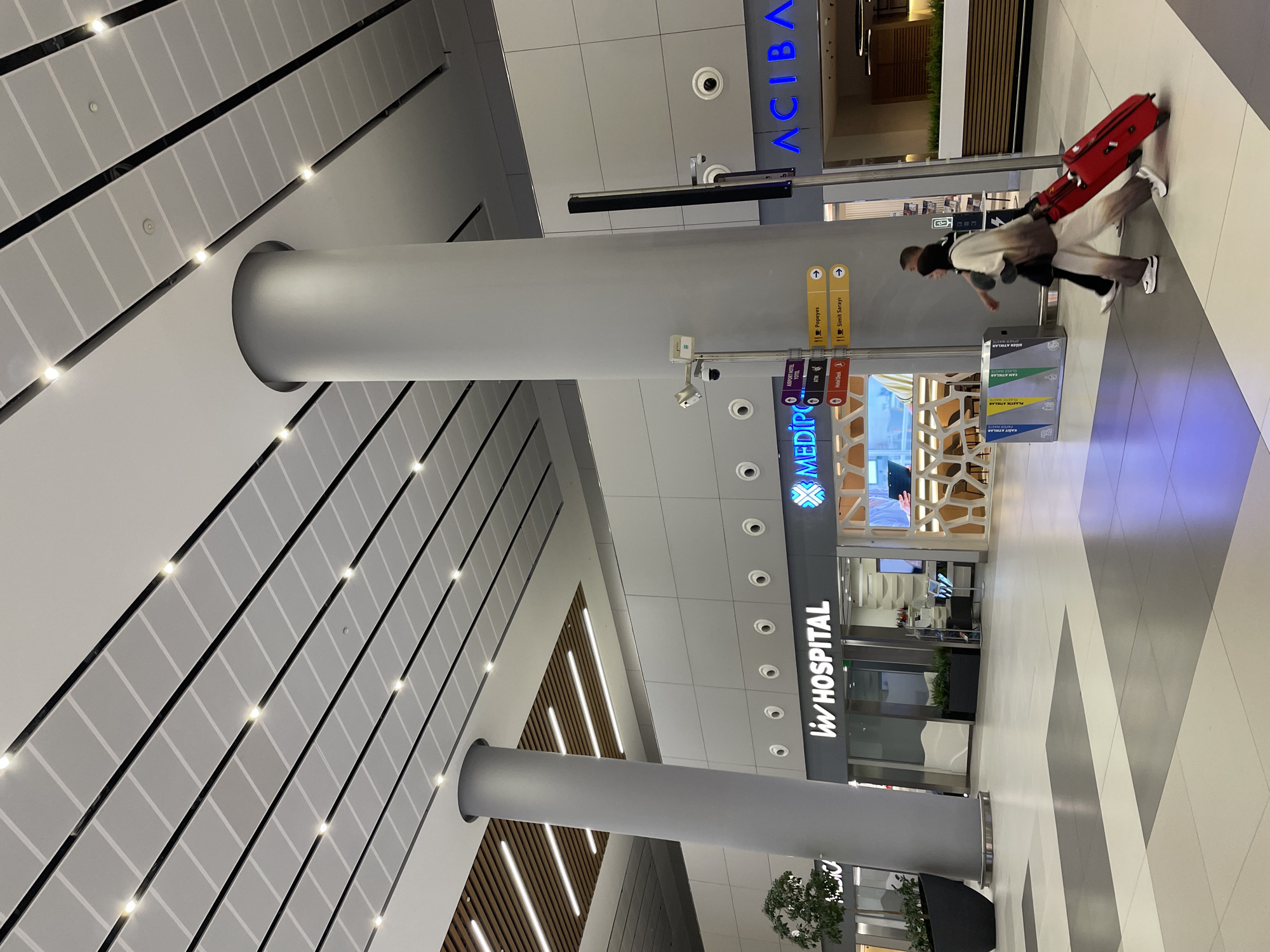 The image captures a contemporary interior space, likely an airport or large public building in Arnavutköy, Turkey, given the presence of airport-related signage. In the foreground, two individuals are walking. The person on the left is a woman wearing a light-colored hooded sweatshirt and pants with a gradient pattern, and is pulling a bright red suitcase. She is walking towards the right side of the frame. The person on the right is partially visible and appears to be wearing dark clothing.
The environment features a modern architectural design with a high ceiling, characterized by a grid of white panels interspersed with recessed lighting and dark linear accents. Large, cylindrical grey columns support the ceiling structure. Several establishments are visible along the wall in the background, including "LIV HOSPITAL" and "ACIBADEM". Directional signage is also present, indicating amenities like "AIRPORT HOTEL", "ATM", "Hotel Desk", "Popeyes", and "Simit Sarayı". A set of recycling bins is positioned near the signage. The floor is tiled in a pattern of light grey and darker grey squares. In the far left of the image, a potted plant in a dark container is visible. The lighting is bright and diffused, creating a well-lit, sterile atmosphere typical of a modern transit hub.
