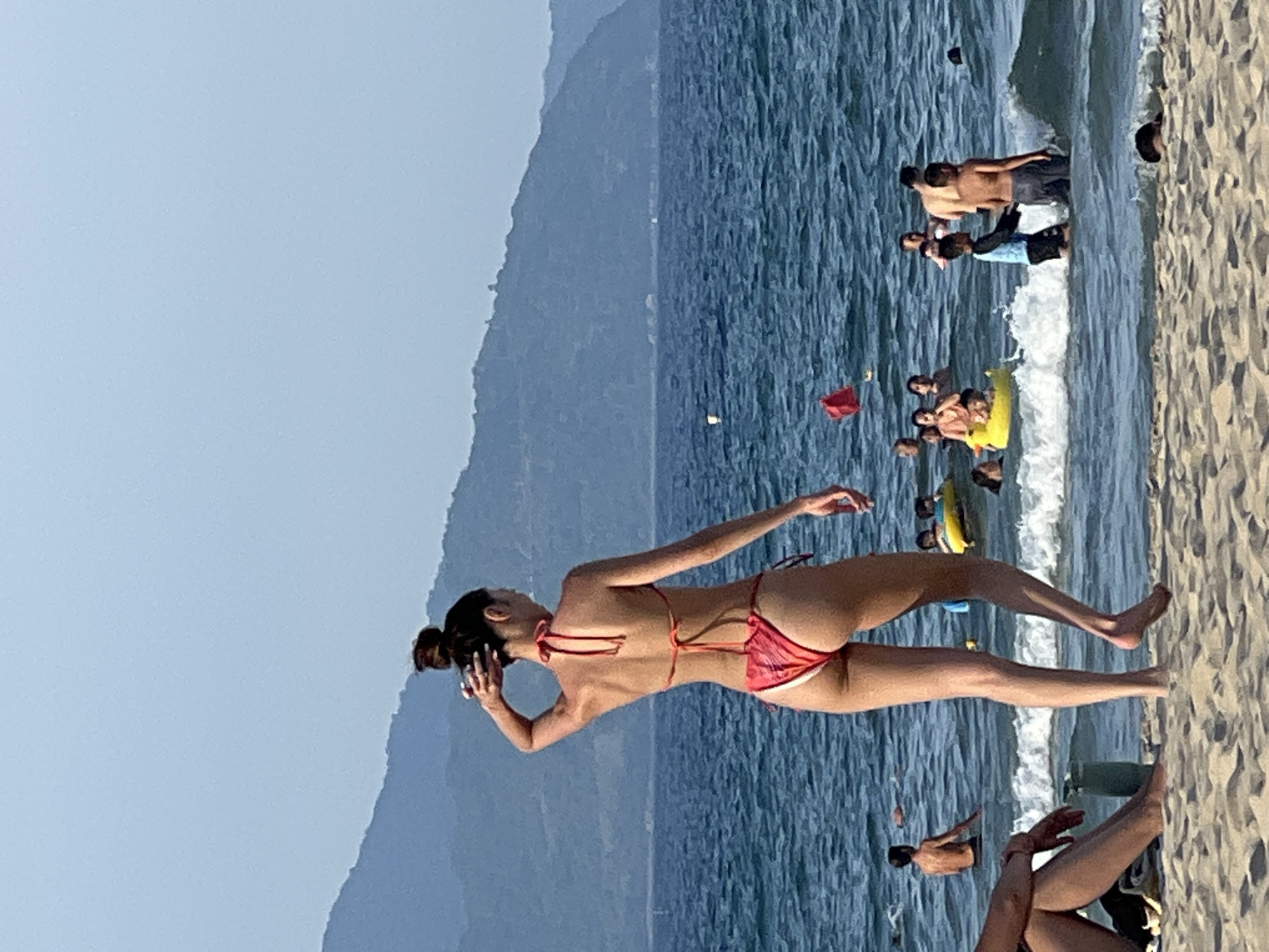 A full shot captures a beach scene in Da Nang, Vietnam, with a woman in a red bikini standing on the sand, facing away from the camera and looking out at the ocean. She has her hand on her head. The ocean is blue with gentle waves, and several people are visible in the water. Some are swimming, while others are playing with inflatable toys. In the background, lush green mountains rise against a clear blue sky. The sand in the foreground is light brown and textured. A person's legs are visible in the bottom left corner of the frame, sitting on the sand.