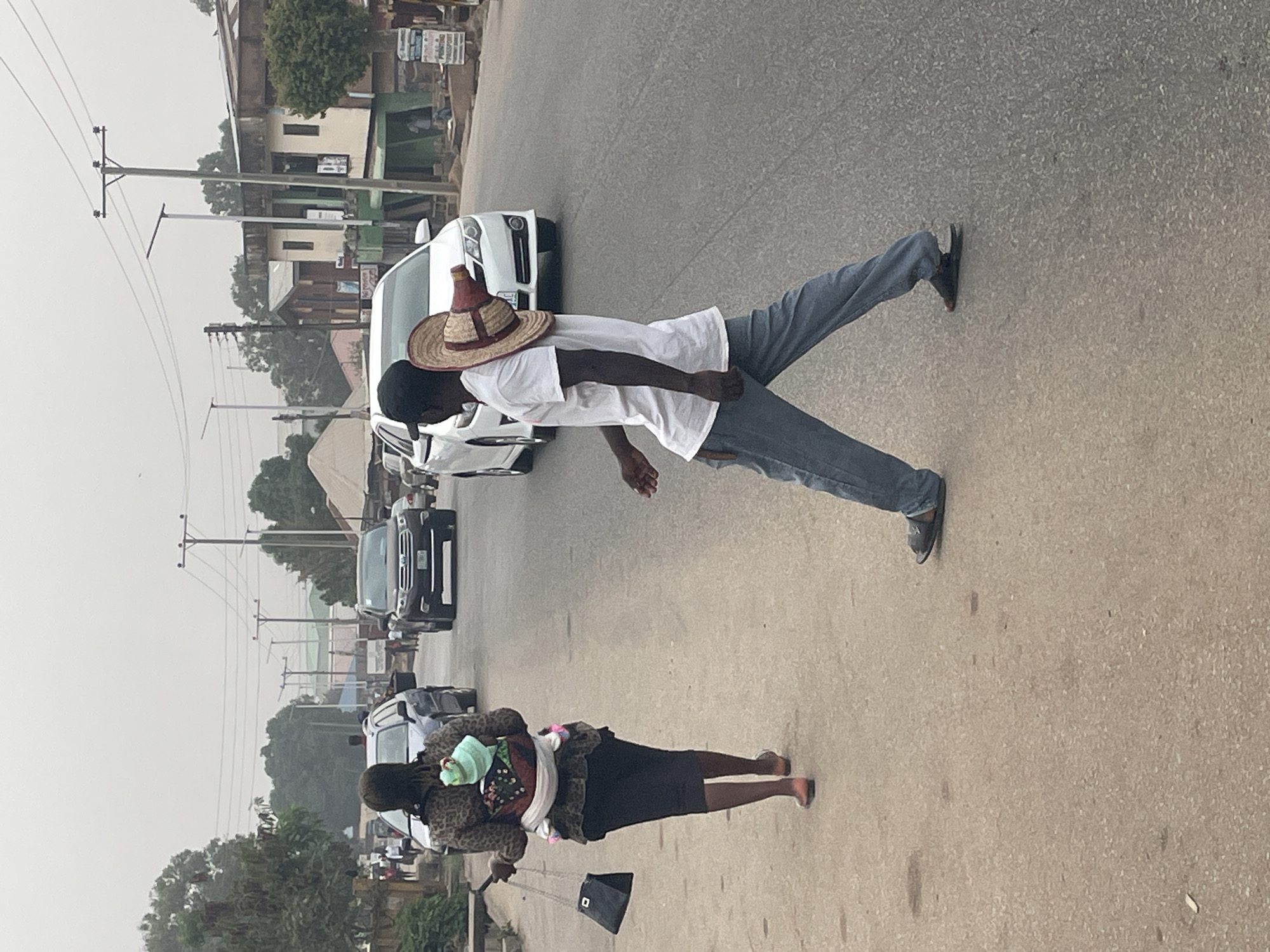 The image, rotated counter-clockwise, captures an outdoor street scene, likely in an urban or semi-urban area consistent with Jos, Nigeria. An asphalt road dominates the lower half, with buildings and utility poles lining the background.
On the right side of the frame, a man with dark skin stands near the road. He wears a white short-sleeved t-shirt, blue jeans, and dark sandals or shoes. A straw hat with red and brown detailing rests on the back of his head. He appears to be looking towards the left of the frame (his right). A white sedan or SUV is partially visible behind him.
Further down the road, closer to the bottom center, a woman with dark skin walks away from the viewer. She is dressed in a black skirt and a patterned jacket, possibly leopard print. A baby, wrapped in a light green cloth, is secured to her body with a white fabric wrap. She carries a black handbag with a chain strap in her left hand, and her feet appear to be bare or in very light sandals.
The street shows signs of activity with several other cars visible in the distance, including a dark SUV. Utility poles with numerous overhead wires run along the side of the road. Buildings, some appearing to be one or two stories with green or yellowish architectural elements, are visible behind the poles. The sky is hazy or overcast, indicating daytime with diffused light. Some text is visible on signs attached to the buildings in the background, but it is too indistinct to be legible. The scene depicts everyday street life with pedestrians and traffic.